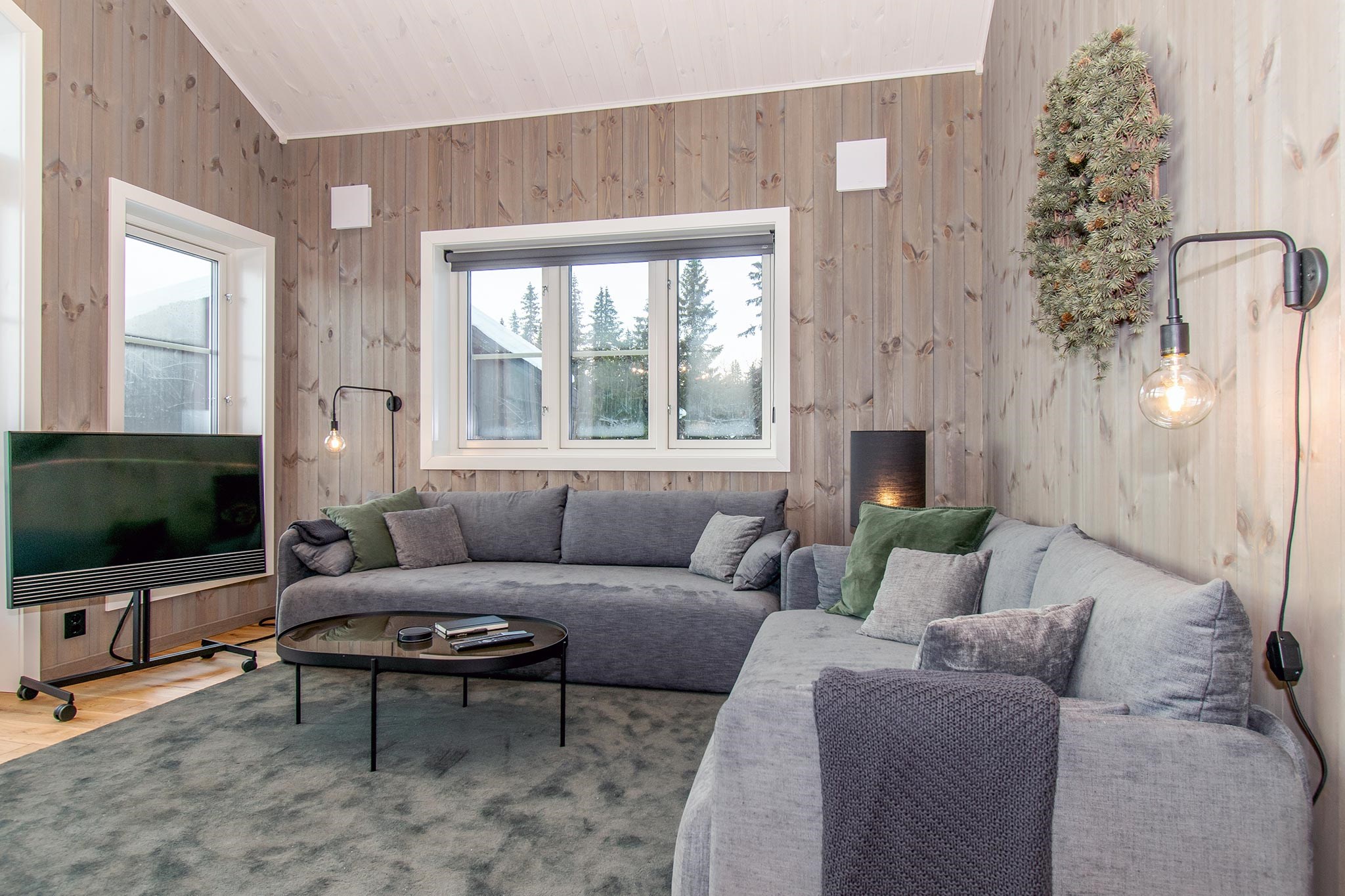Moderne stue med træpaneler på væggene, to grå sofaer, grønne og grå puder, et rundt sort sofabord, et fladskærms-tv, vægmonterede lamper, en krans og store vinduer, der lukker naturligt lys ind.