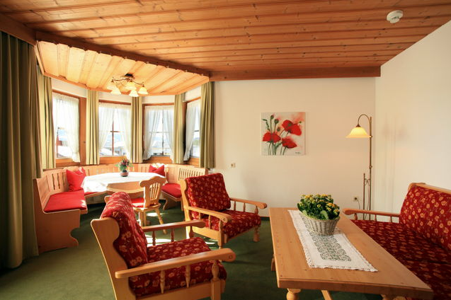 En hyggelig stue med træloft, røde polstrede stole, et træbord med et blomsterarrangement og en hjørnehjørne med karnappvinduer, grønne gardiner og naturligt lys. Et maleri hænger på den hvide væg.