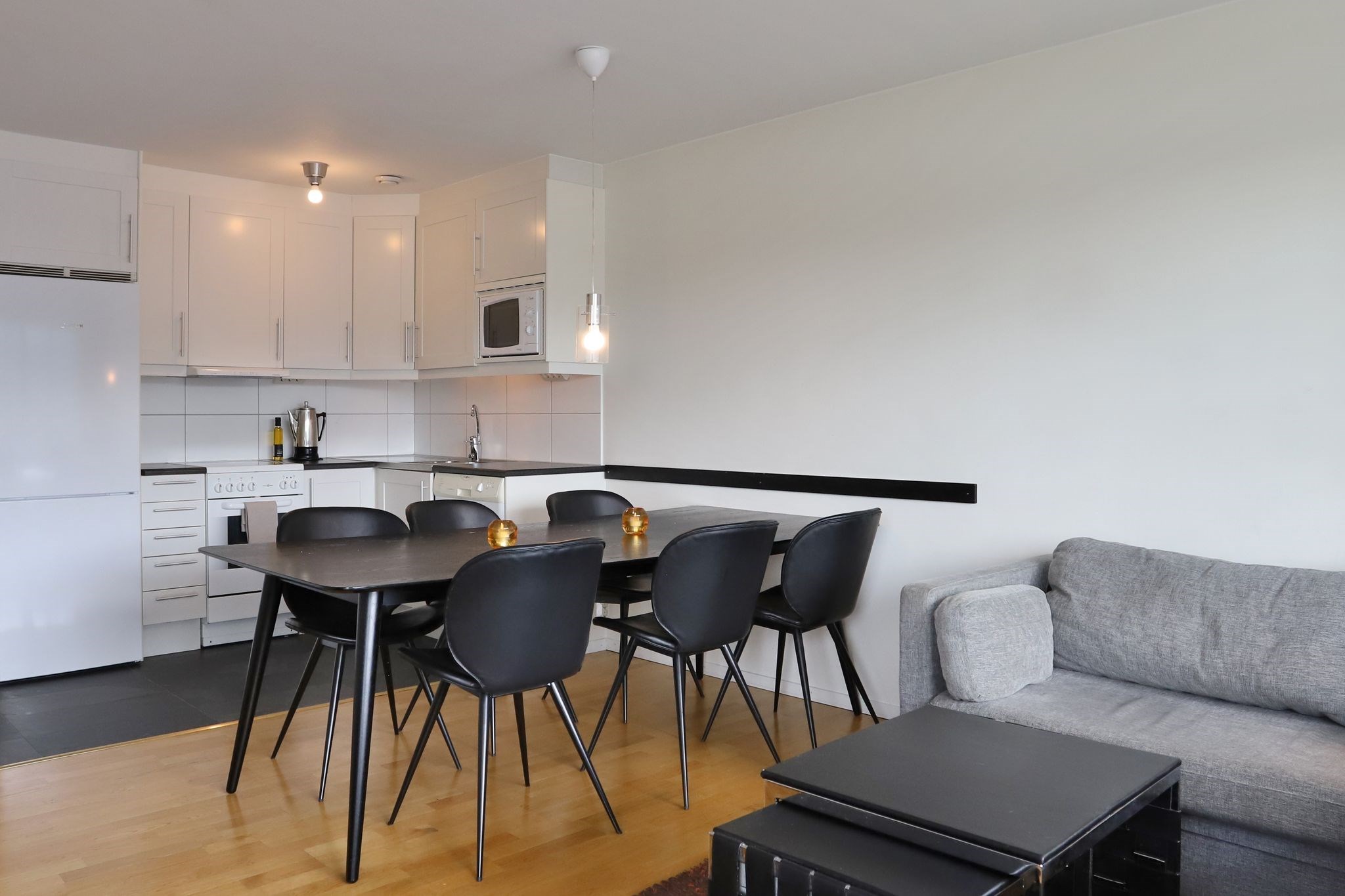 Moderne åbent køkken og spisestue med hvide skabe, sort spisebord og seks sorte stole, grå sofa, trægulv og minimalistisk indretning.