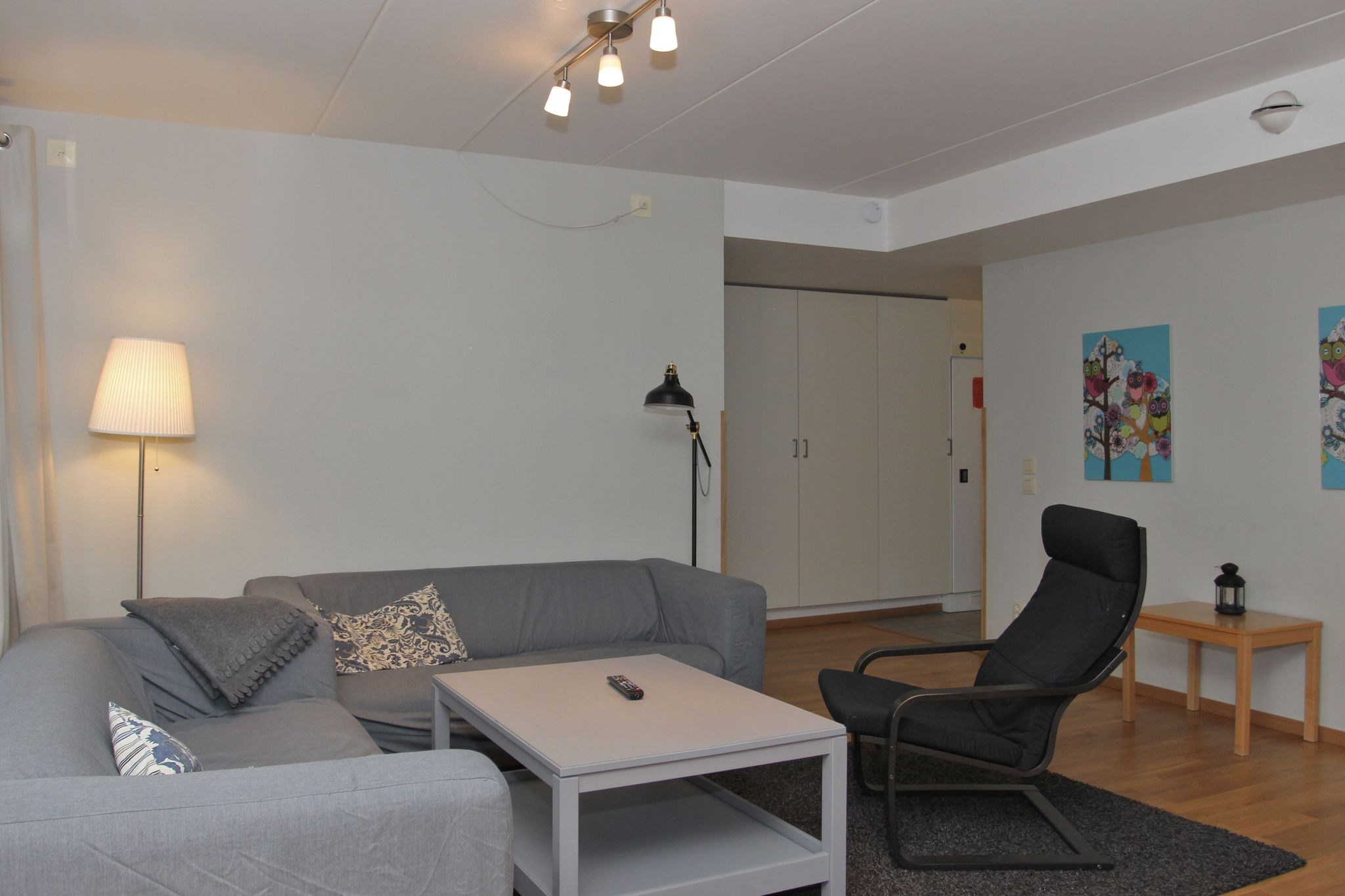 En hyggelig stue med en grå sofa, en sort lænestol, et lysegråt sofabord og en gulvlampe. Kunsttryk hænger på væggen, og i baggrunden er der et klædeskab. En fjernbetjening ligger på sofabordet.