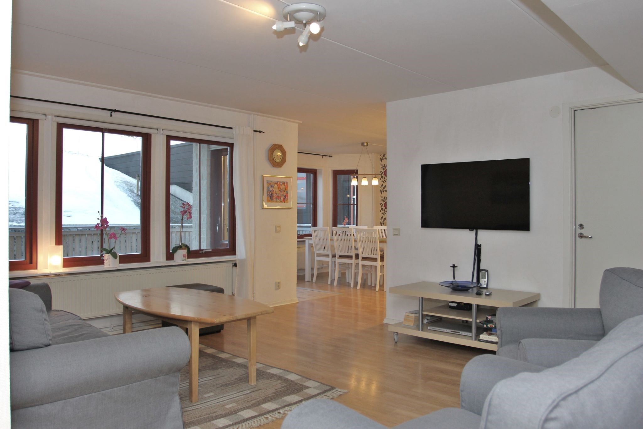 En moderne stue med grå sofaer, et sofabord i træ, et stort vægmonteret tv og store vinduer med udsigt over sneen. Rummet åbner ind i en spisestue med et hvidt bord og stole.