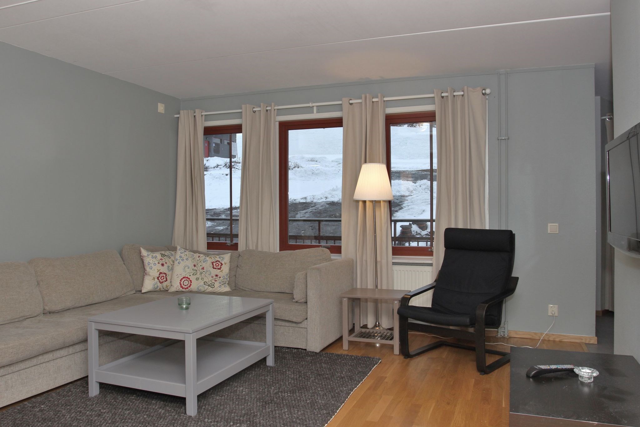 En moderne stue med en grå hjørnesofa, mønstrede puder, en sort lænestol, et gråt sofabord, en gulvlampe og store vinduer med beige gardiner, der viser et snedækket udendørssceneri.