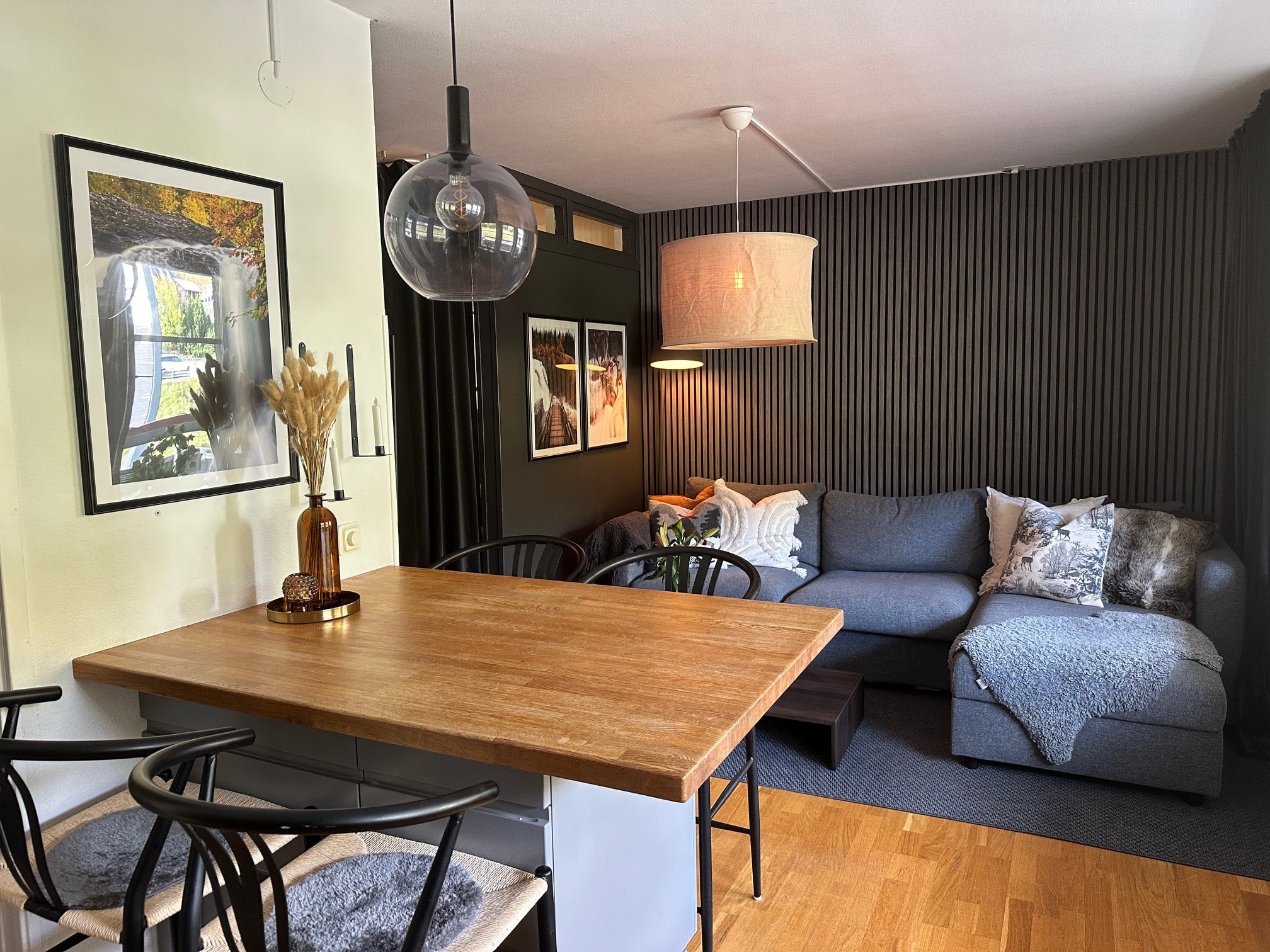 En hyggelig stue med et spisebord i træ, sorte stole, en grå sofa med puder og tæpper, moderne pendellamper, indrammet vægkunst og mørke vægpaneler med tekstur.