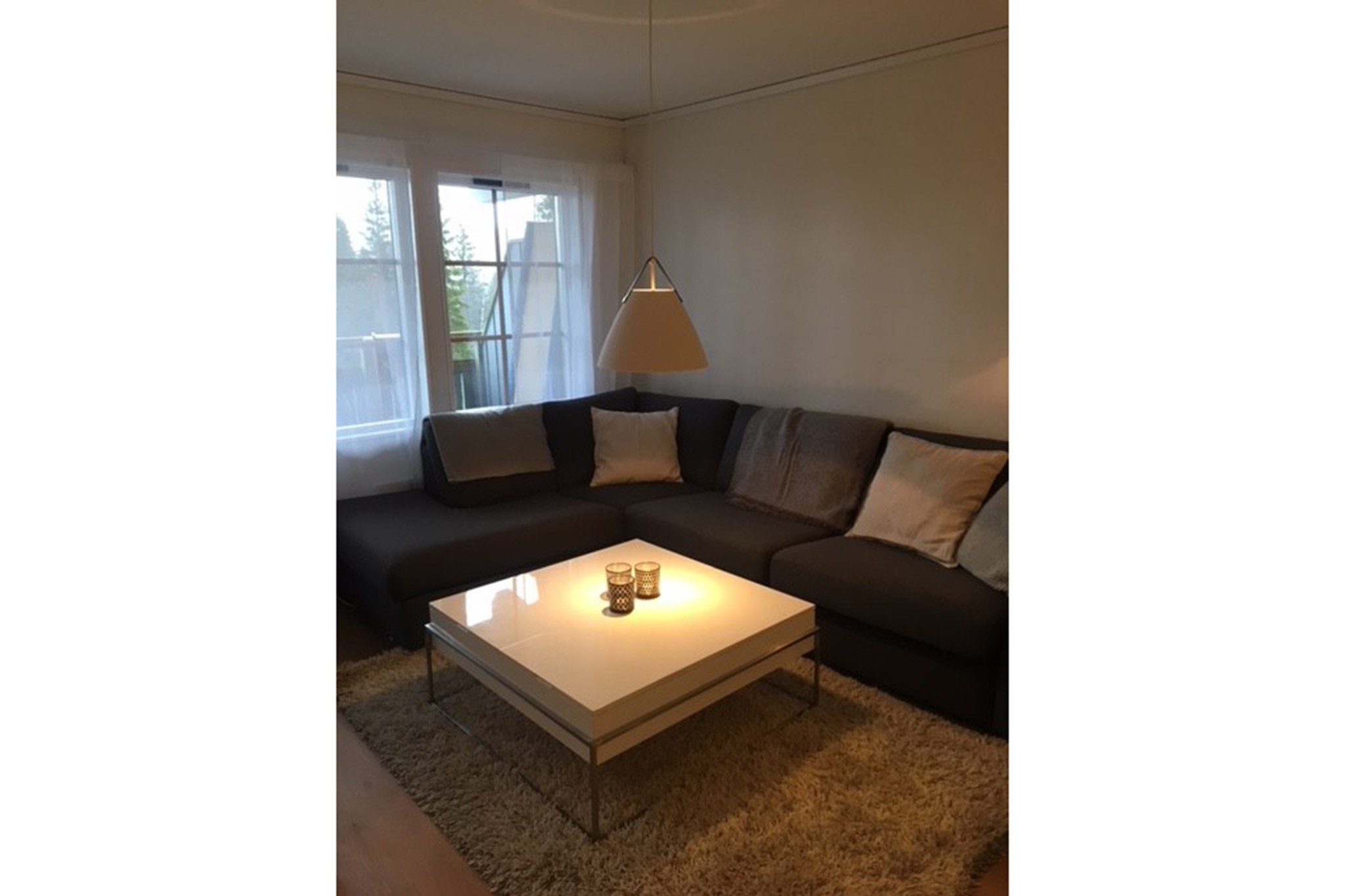 En hyggelig stue med en mørk sofa, lyse puder, et hvidt sofabord med stearinlys, et beige tæppe og store vinduer med tynde gardiner, der lukker naturligt lys ind.