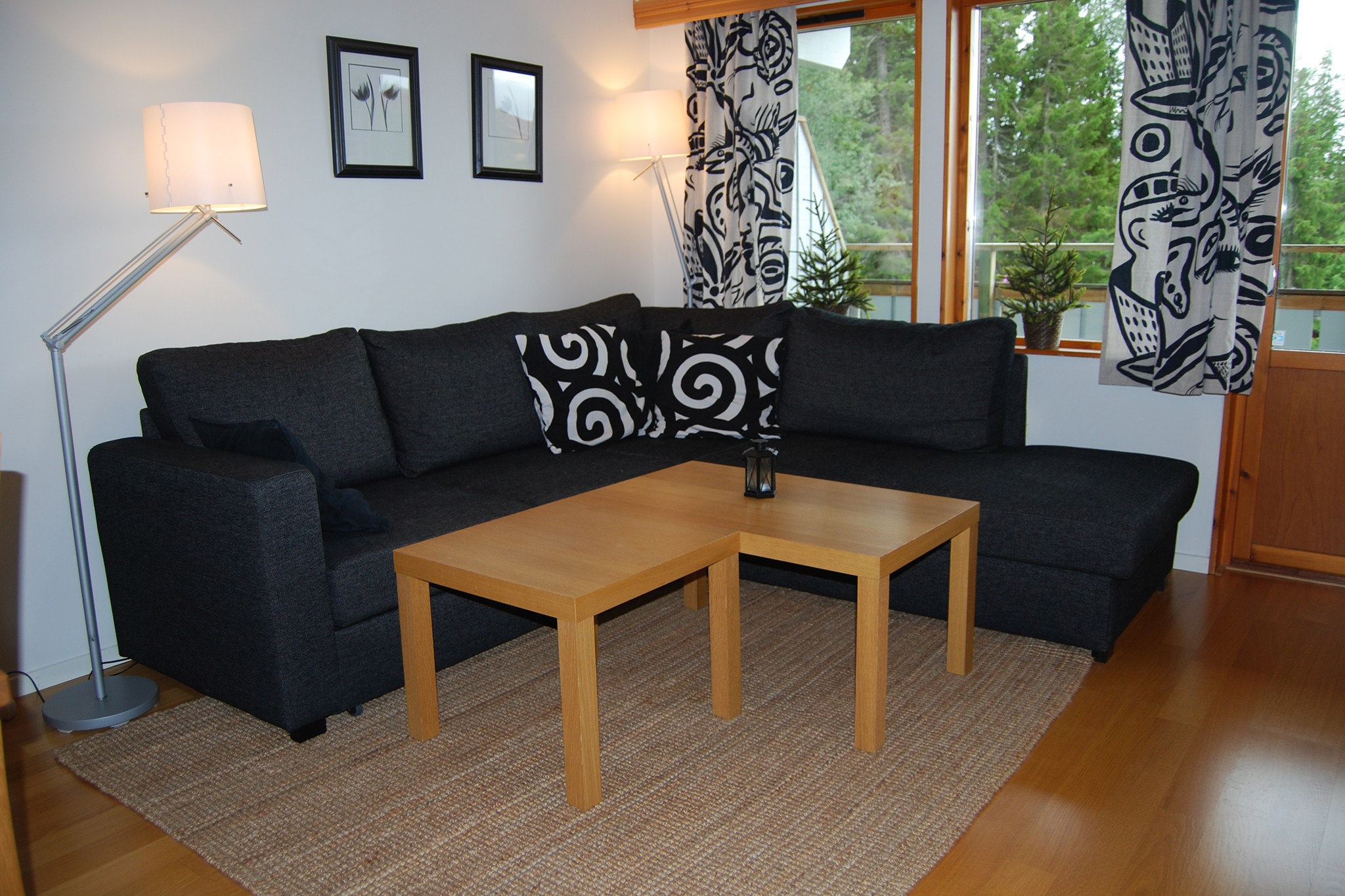 En moderne stue med en mørkegrå hjørnesofa, sort-hvide mønstrede puder, et lyst træsofabord, en gulv- og væglampe, indrammet kunst og store vinduer med mønstrede gardiner med udsigt over grønne områder.