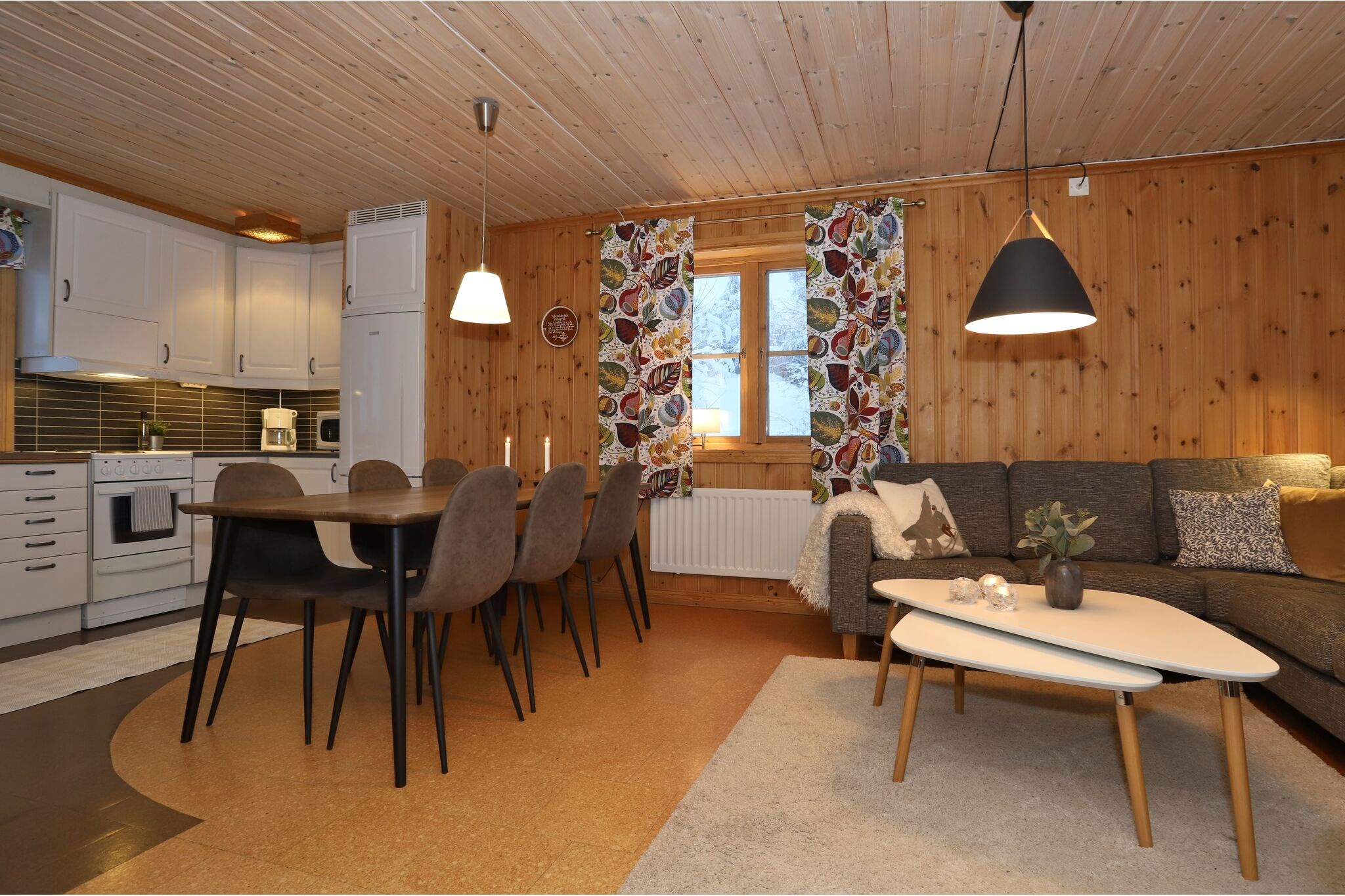 En hyggelig opholds- og spisestue i træ med et moderne køkken, langt spisebord med otte stole, grå sektionssofa, farverige gardiner, pendellamper og sofaborde på et beige tæppe.