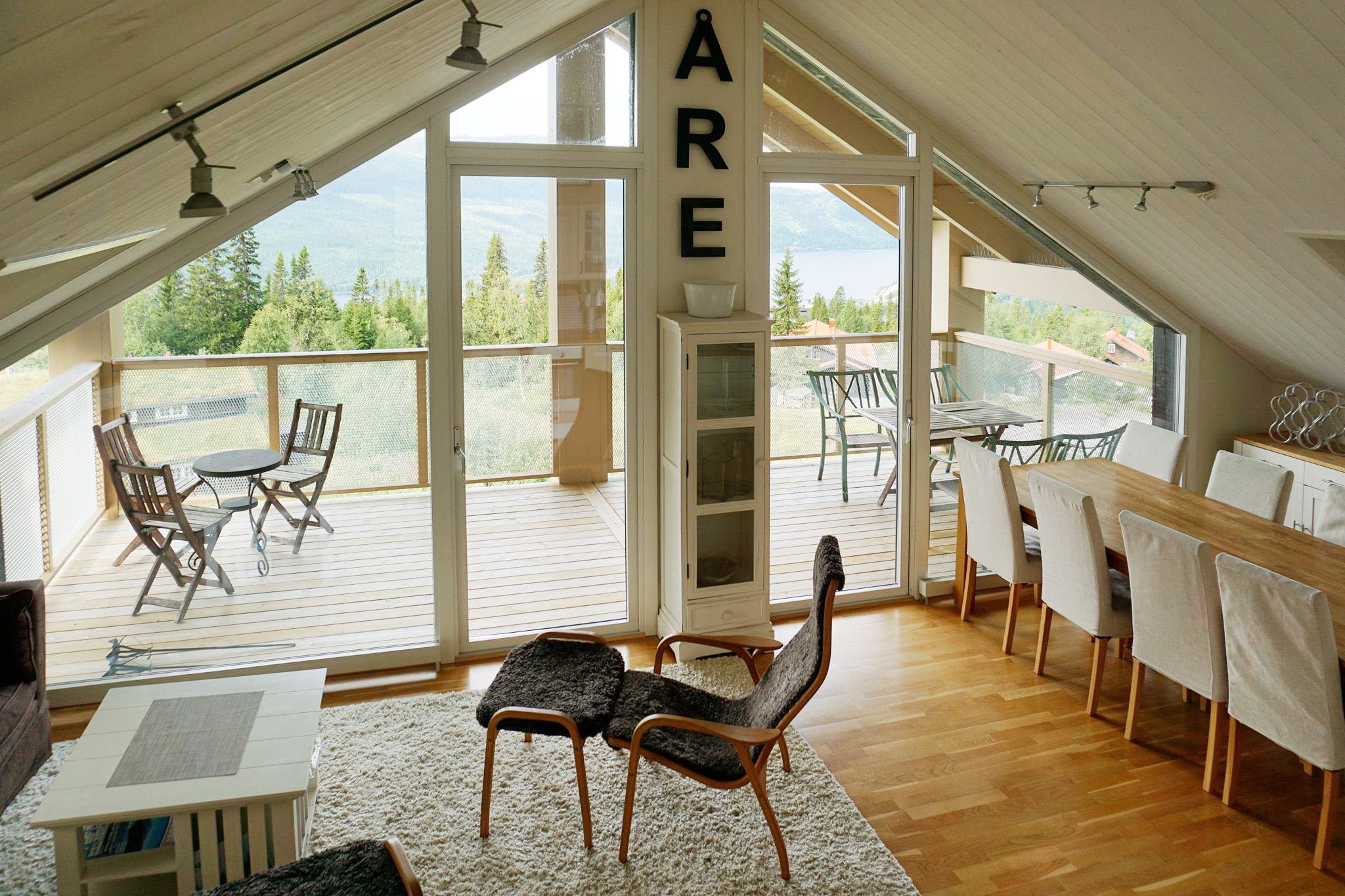 En hyggelig, moderne opholds- og spisestue på loftet med trægulve, en lænestol, et spisebord med hvide stole og store vinduer, der åbner ud til en balkon med udsigt over bjergene og skoven. Ordet ÅRE vises lodret på en søjle.