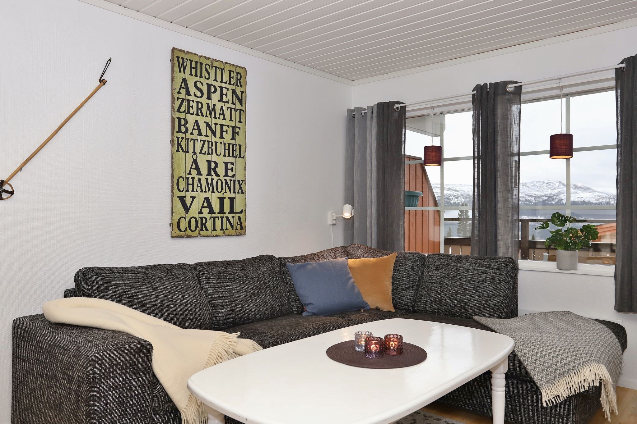 En hyggelig stue med en mørkegrå sofa, tæpper, et hvidt sofabord med stearinlys, vægkunst med en liste over skisportssteder, grå gardiner og store vinduer, der viser det sneklædte bjerglandskab udenfor.