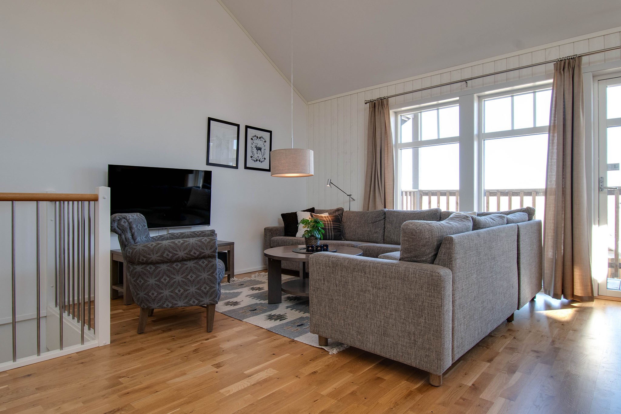 Lys stue med store vinduer, grå hjørnesofa, mønstret lænestol, rundt sofabord, TV på et bord, lyst trægulv, beige gardiner og indrammet kunst på hvide vægge.