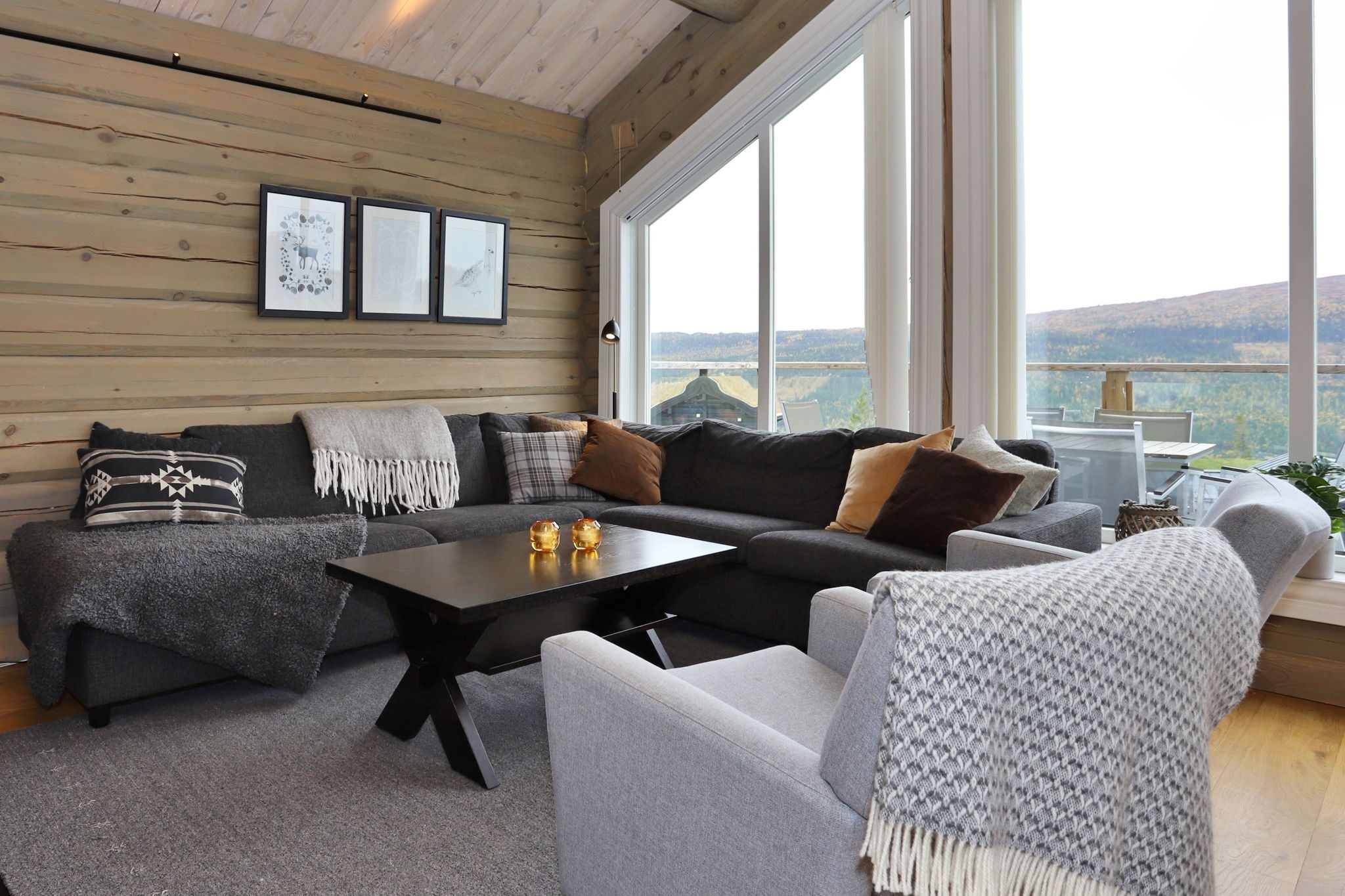 En hyggelig stue med en grå sofa, mønstrede og ensfarvede hynder, et sofabord med stearinlys, indrammet vægkunst, store vinduer med en smuk udsigt og bløde tæpper draperet over møblerne.