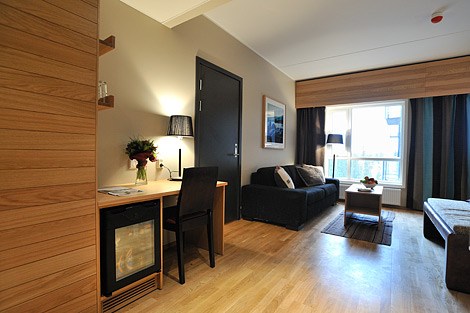 Moderne hotelværelse med trægulve, et skrivebord med stol og lampe, et minikøleskab, en sofa, et stort vindue med gardiner og en seng, der delvist er synlig til højre. Et indrammet billede hænger på væggen.