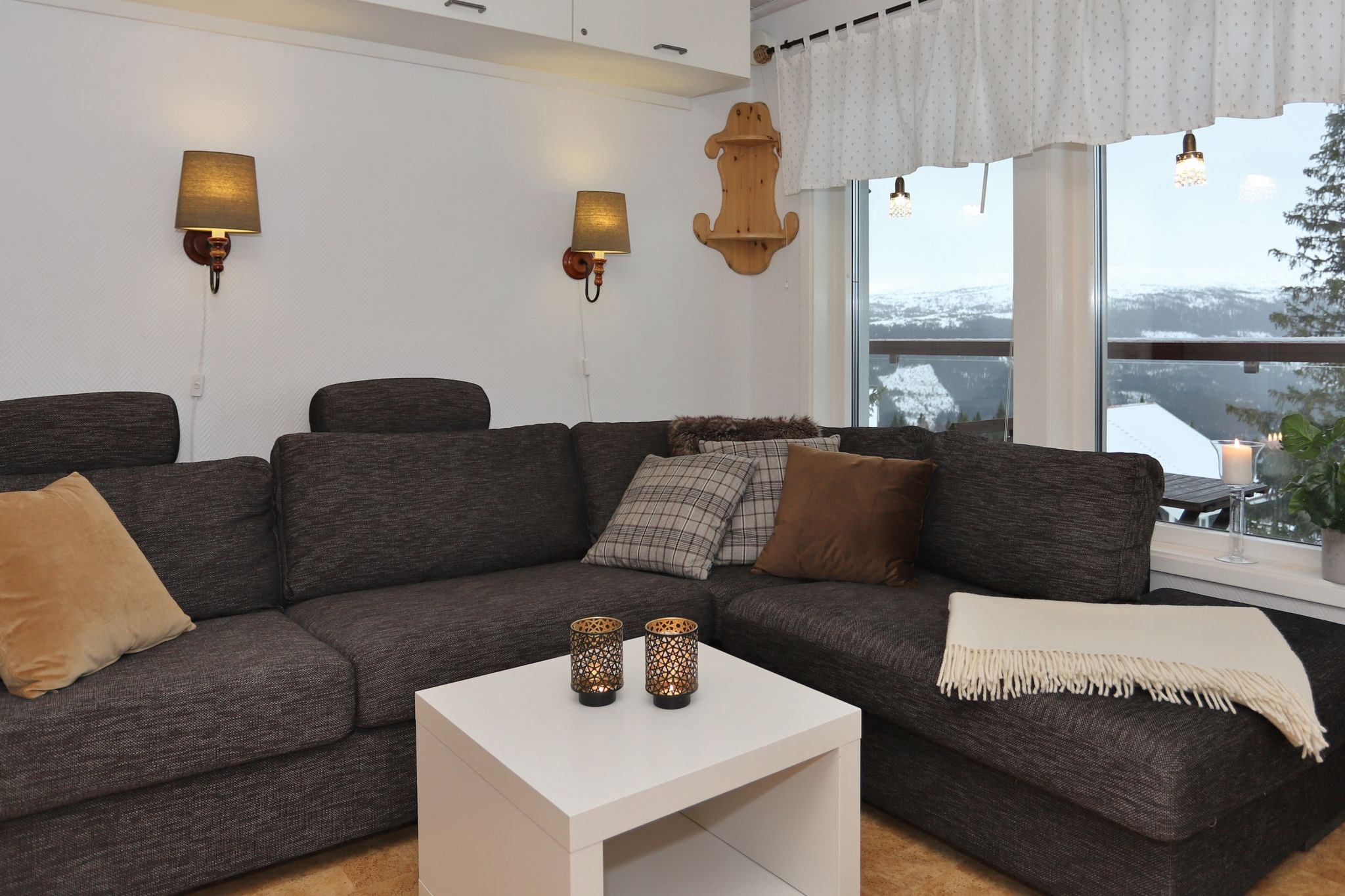 En hyggelig stue med en mørkegrå sofa, forskellige puder, et hvidt sofabord med tre stearinlys, væglamper og et stort vindue med udsigt over bjergene og det sneklædte landskab udenfor.