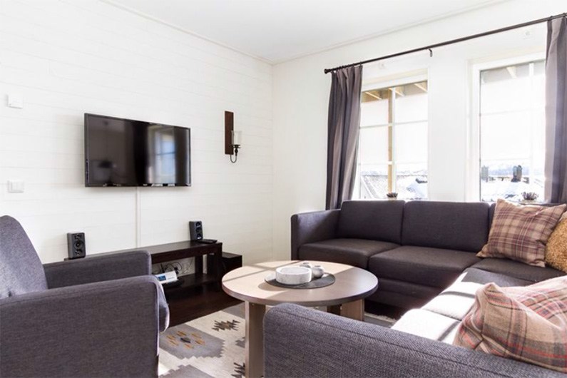 En lys stue med en mørkegrå sofa, en grå lænestol, et rundt sofabord, et tv monteret på en hvid væg, et mediebord og store vinduer med mørke gardiner, der lukker naturligt lys ind.