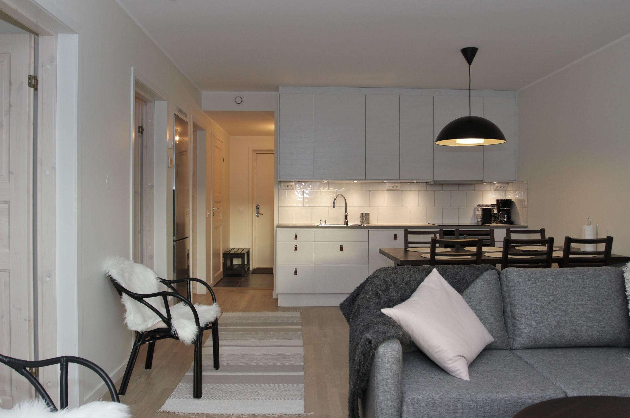 En moderne, minimalistisk stue og køkken med hvide vægge, lyse trægulve, en grå sofa med en lyserød pude, sorte stole, et spisebord og elegante hvide skabe under stærk belysning.