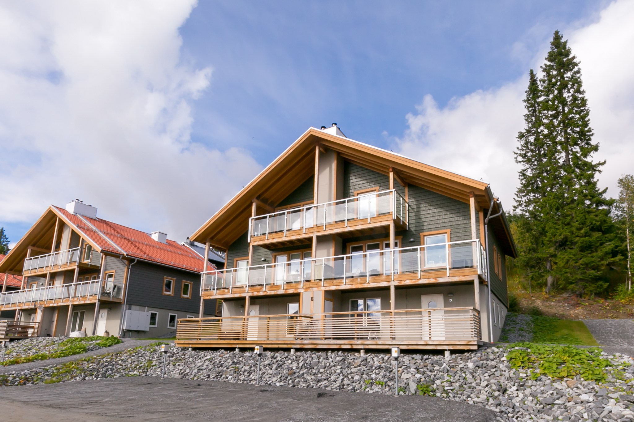 Et moderne, toetagers træhus med rummelige altaner og store vinduer, beliggende på en klippefyldt skråning med et andet lignende hus i nærheden; omgivet af træer under en delvist overskyet himmel.
