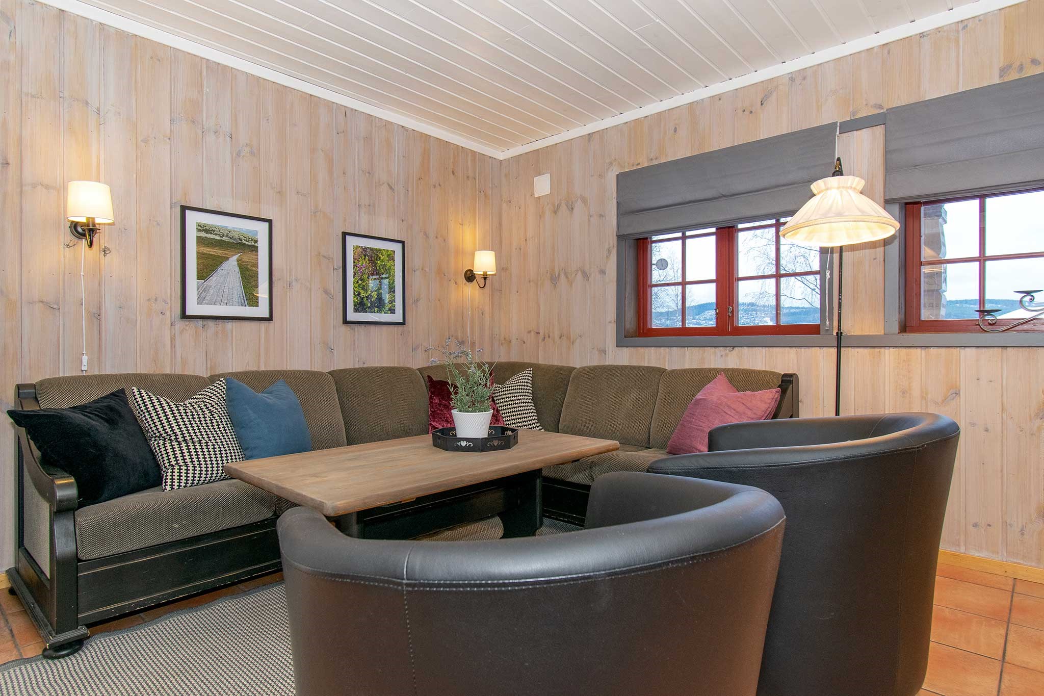En hyggelig stue med lyse træpaneler på væggene, en hjørnesofa med forskellige puder, to sorte lænestole, et sofabord i træ, indrammet vægkunst og store vinduer med grå romerske gardiner.