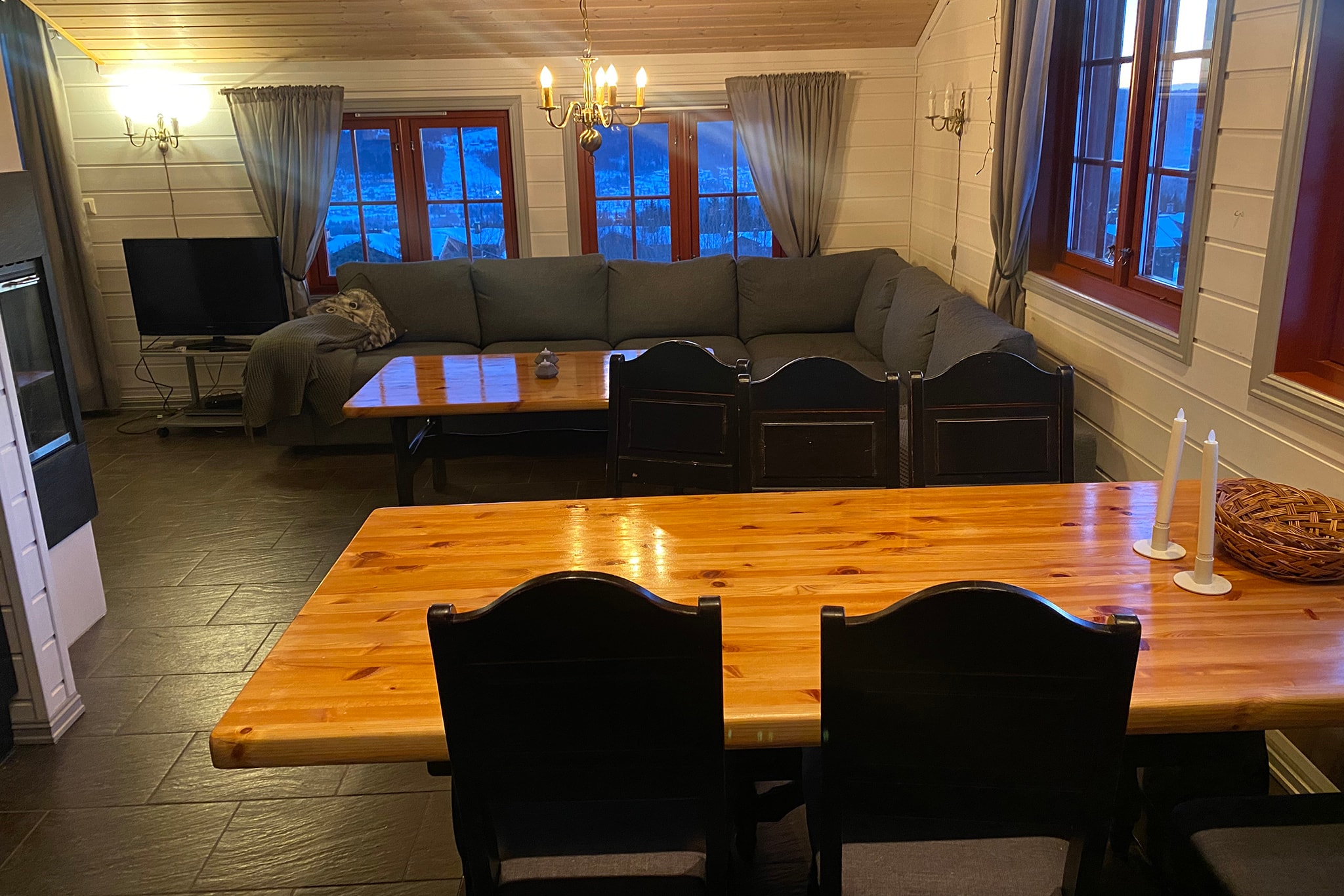 Hyggelig stue med en stor grå sofa, TV, spisebord i træ med mørke stole og vinduer med gardiner, der viser udsigten til sneen udendørs. Varm belysning og træloft skaber en indbydende atmosfære.