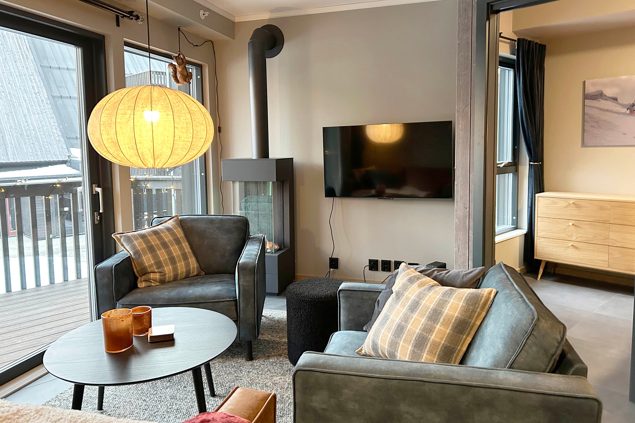 En moderne stue med to grå lænestole, et rundt sofabord, et vægmonteret tv, en brændeovn og en stor papirlanterne. Vinduer fra gulv til loft og en dør åbner ud til en balkon.