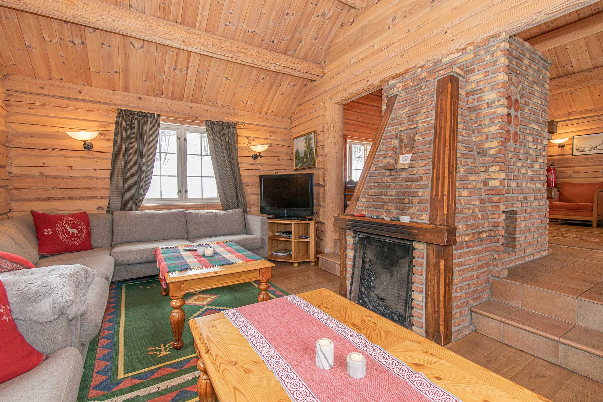 En hyggelig stue i bjælkehytte med trævægge og -loft, en pejs af mursten, to grå sofaer, et tv og et farverigt mønstret tæppe. Sollys kommer ind gennem to vinduer med gardiner bag sofaerne.