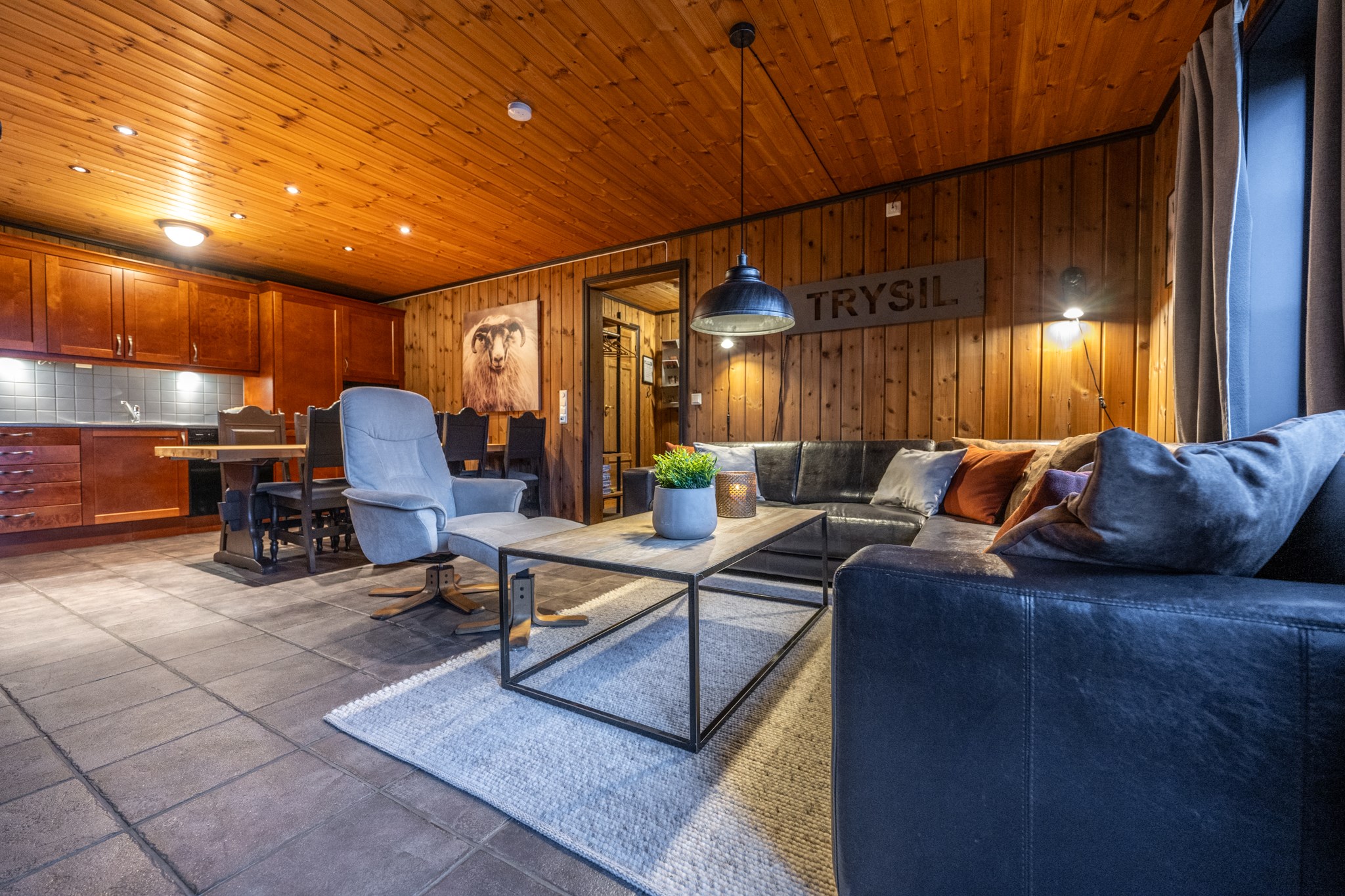 Hyggelig stue med trævægge og -loft, sort lædersofa, moderne lænestol, sofabord, gråt tæppe, spiseplads og et køkken i baggrunden. Varm belysning skaber en indbydende atmosfære.