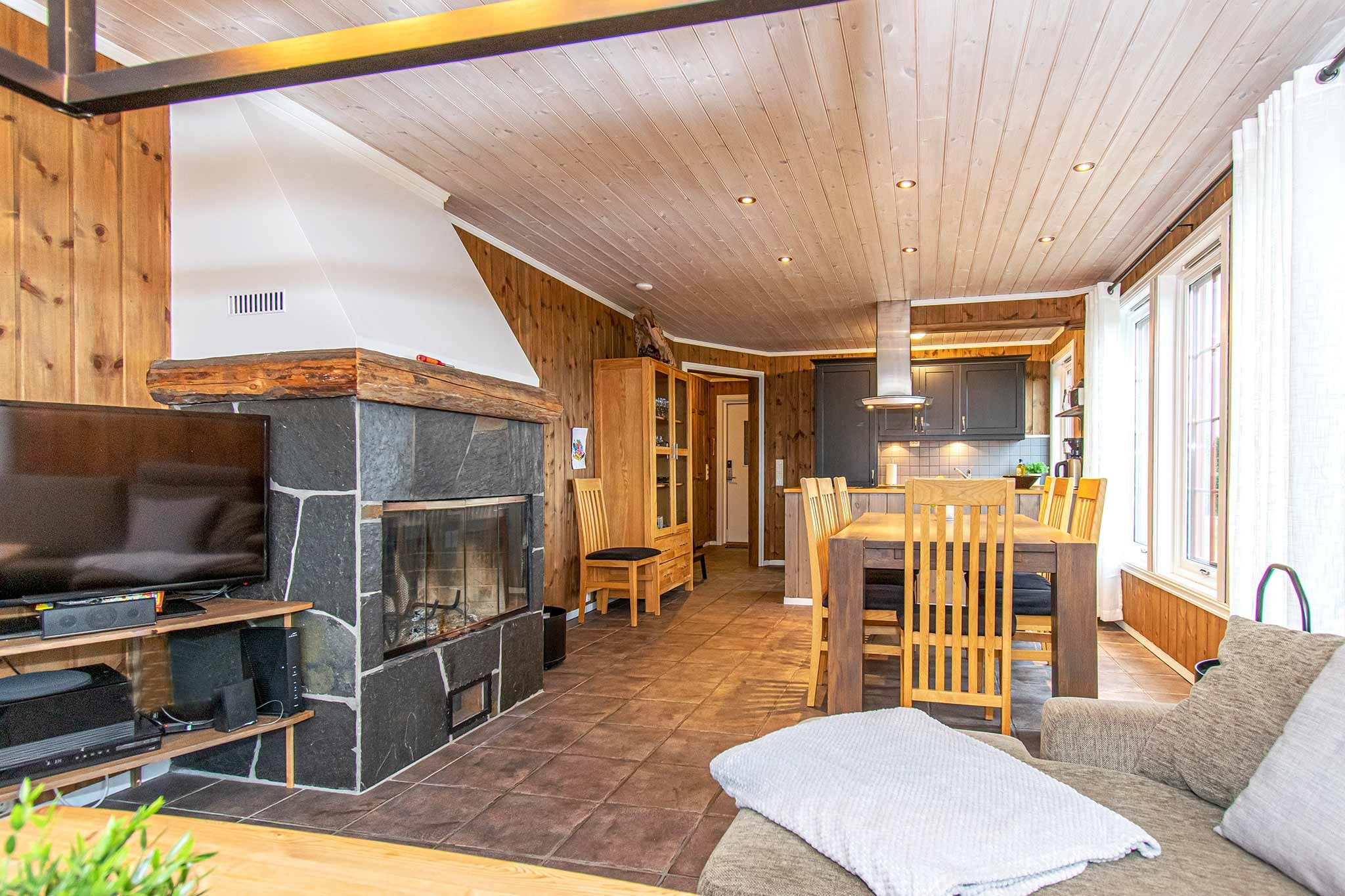 En hyggelig opholds- og spisestue med trævægge og -loft, en stenpejs, et tv, et spisebord med stole, et moderne køkken og store vinduer, der lukker naturligt lys ind.