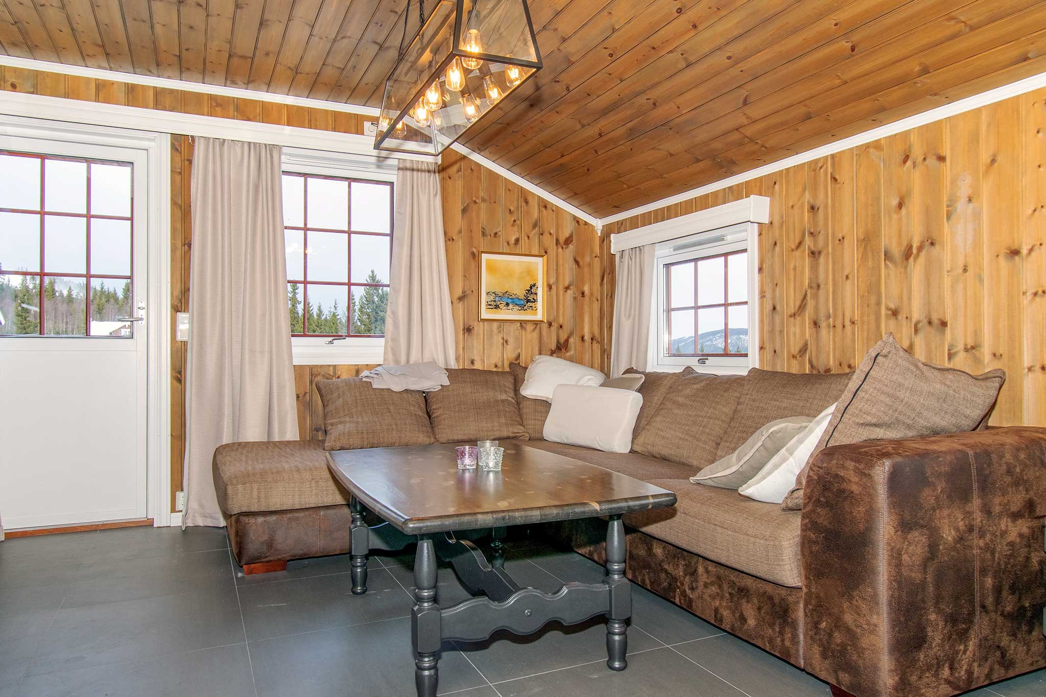 Hyggelig stue i træ med en brun hjørnesofa, puder, et mørkt sofabord i træ og vinduer med beige gardiner. En moderne lampe hænger fra loftet med træpaneler.