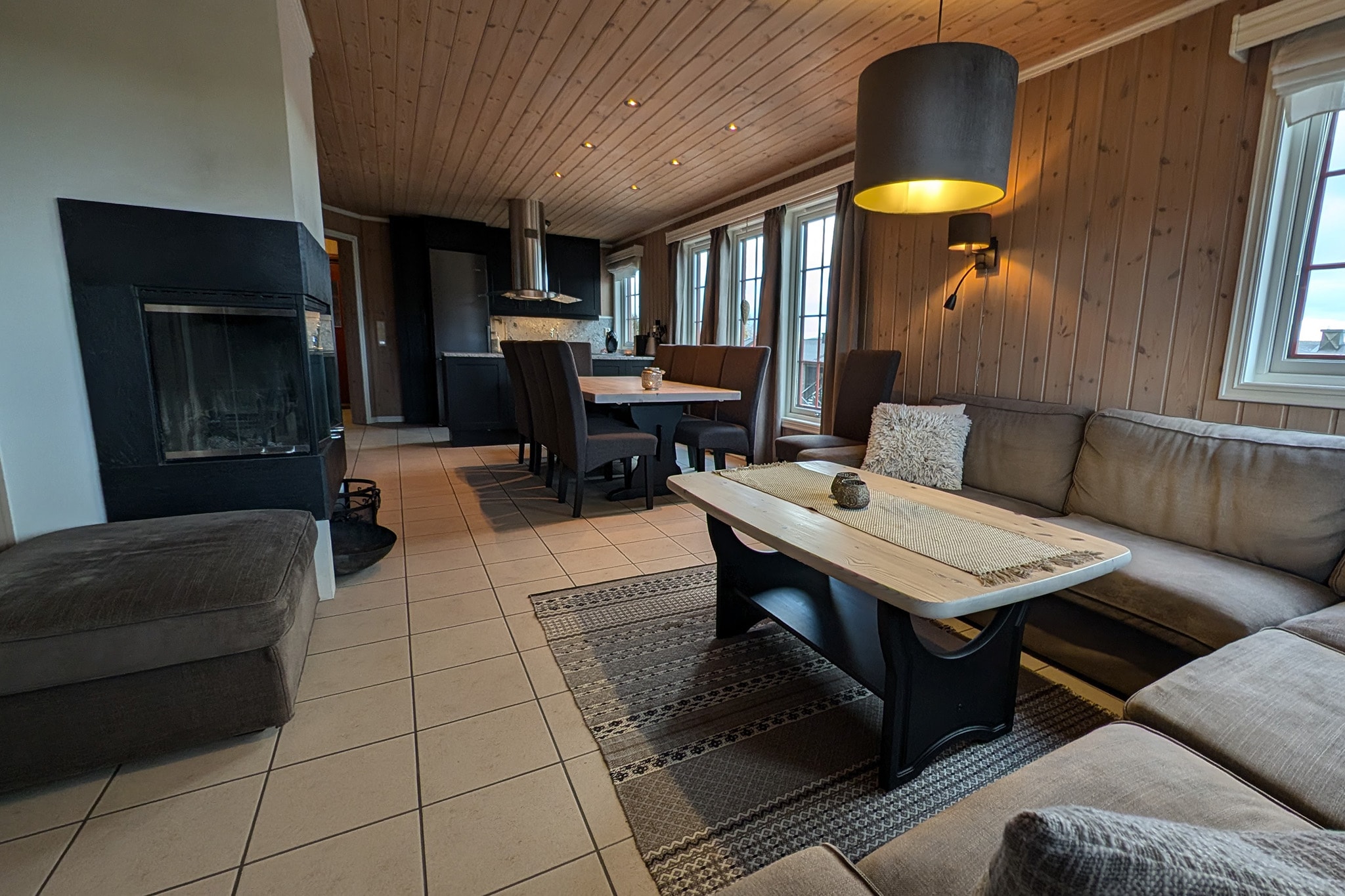 En hyggelig stue med en sofagruppe, et sofabord i træ og en pejs. Rummet har træpaneler på væggene, flisebelagt gulv, spiseplads med bord og stole samt store vinduer, der lukker naturligt lys ind.