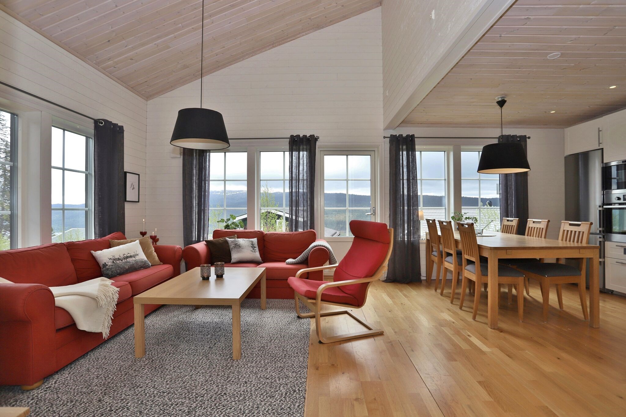 Moderne opholds- og spisestue med trægulve, røde sofaer, en rød lænestol, lyse træmøbler, store vinduer med tynde gardiner og en smuk udsigt udad. Værelset er lyst med neutrale og røde accenter.
