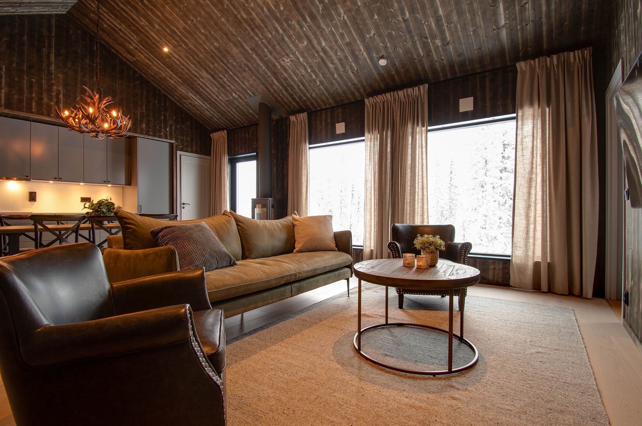 En hyggelig stue med en beige sofa, to mørke læderlænestole, et rundt sofabord i træ og store vinduer med beige gardiner, der lukker naturligt lys ind. Loftet og væggene er i mørkt træ, hvilket giver en varm, rustik stemning.