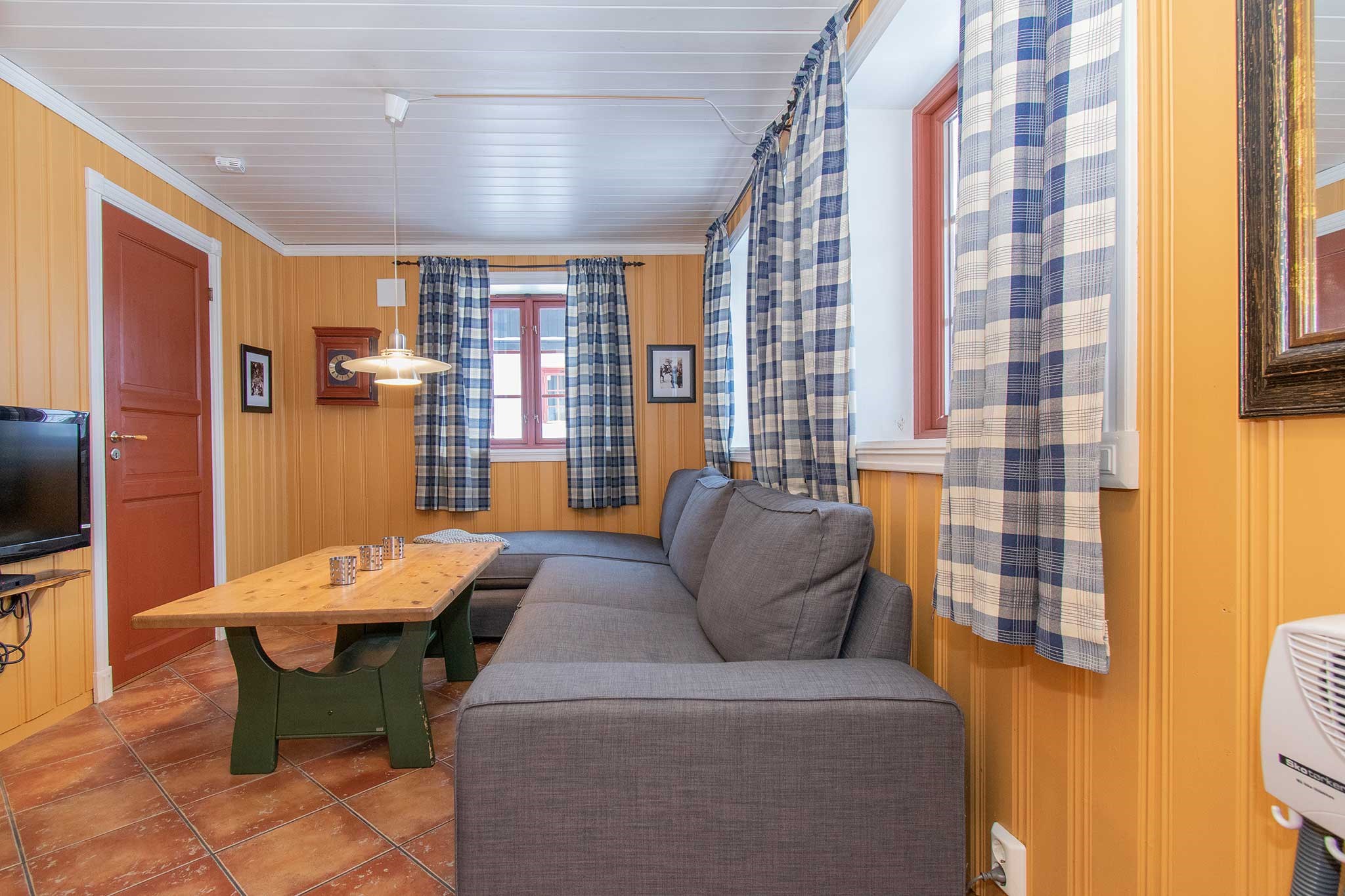 En hyggelig stue med gule trævægge, blå ternede gardiner, en grå hjørnesofa, et sofabord i træ med grønne ben og et tv-bord på et stativ. Gulvet er flisebelagt i brune og orange toner.