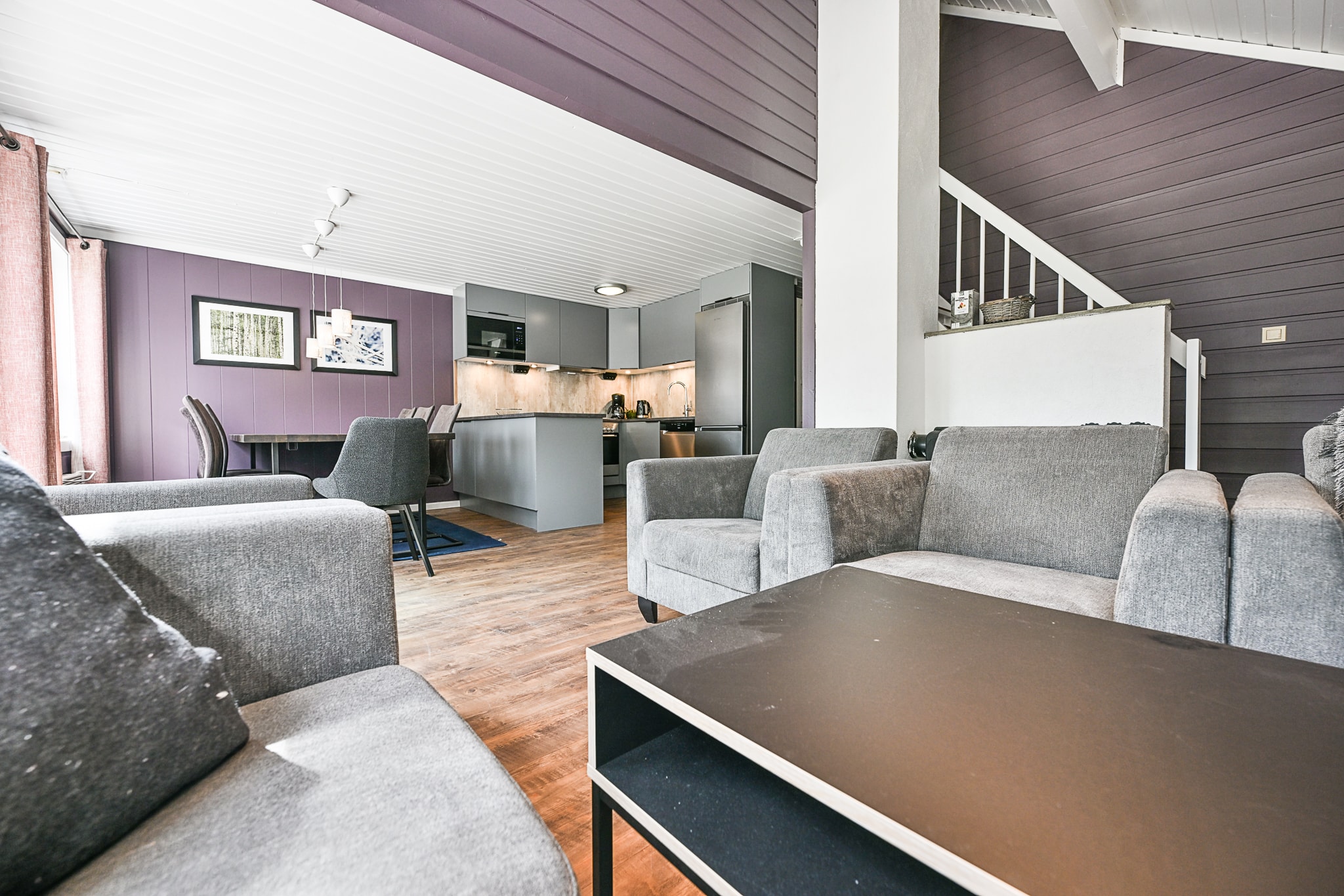 Moderne åben stue med grå sofaer, sort sofabord, spisebord og et elegant køkken. Lyse trægulve og lilla-grå vægge skaber en hyggelig, moderne atmosfære. Trappe fører til en øvre etage.