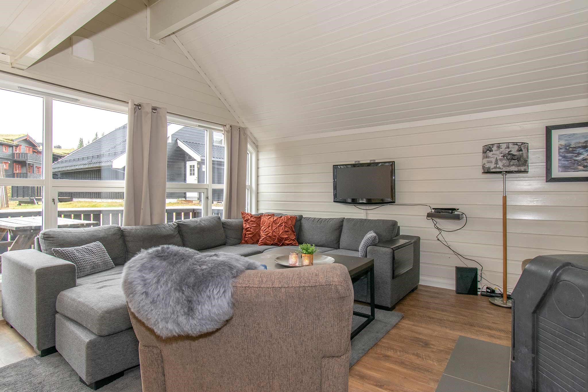 En hyggelig stue med en grå sofa, orange og grå hynder, et sofabord, vægmonteret TV og store vinduer, der lukker naturligt lys ind. Værelset har hvide trævægge og -loft samt trægulv.
