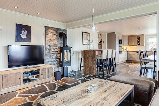 Hyggelig åben stue med rustikke træmøbler, tv, brændeovn på en stenmur og køkken med morgenmadsbar og taburetter i baggrunden. Stenmønstret gulv i hele rummet.