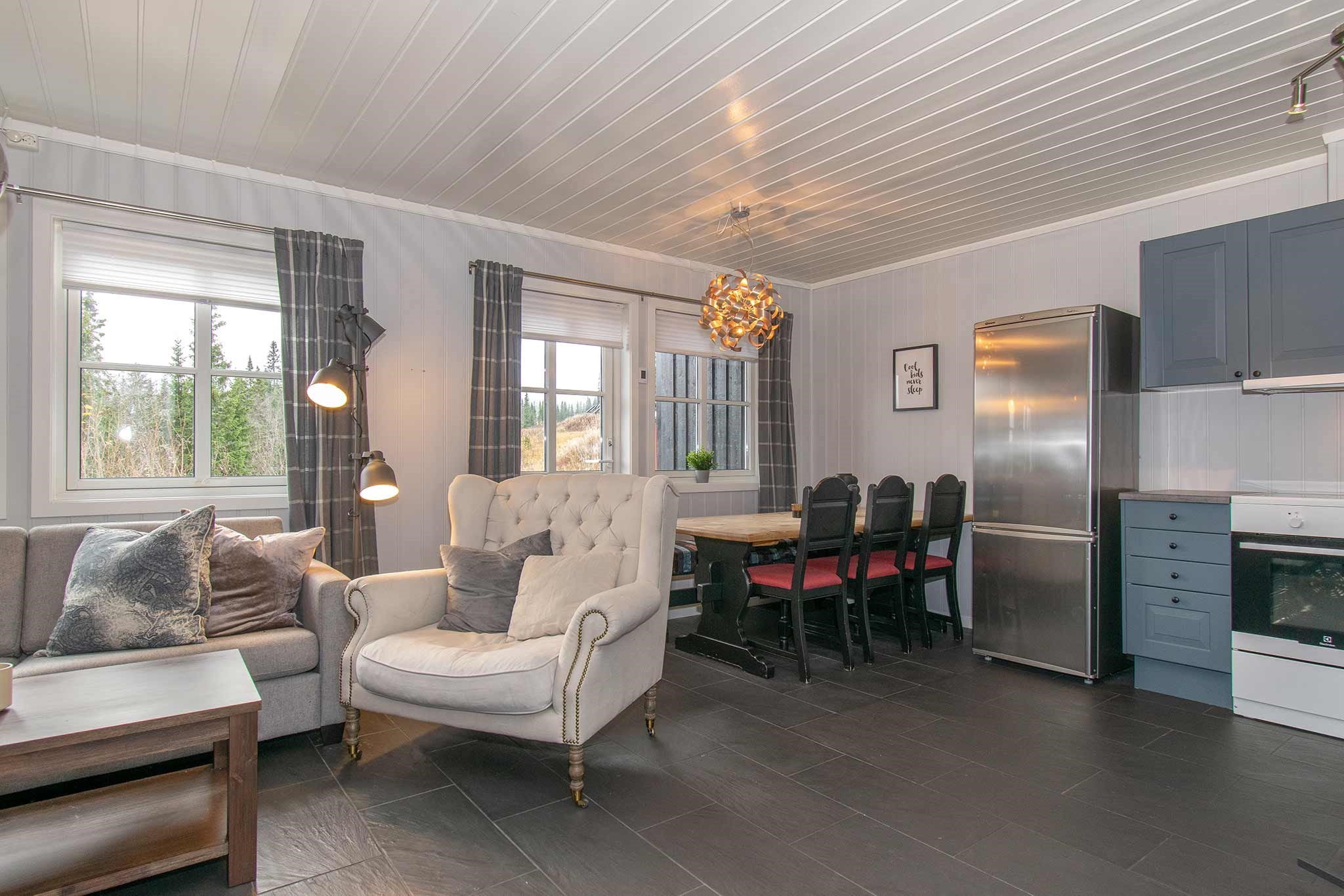 Hyggelig opholds- og spisestue med en grå sofa, beige lænestol, træbord med sorte stole, moderne lampe, blå køkkenskabe, køleskab i rustfrit stål og store vinduer med ternede gardiner.