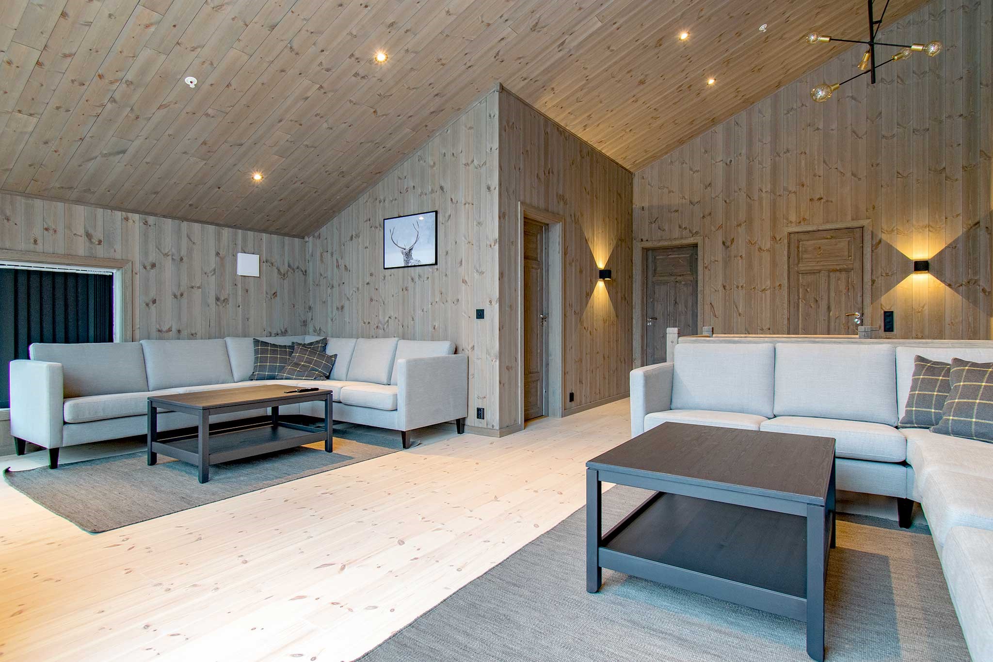 Moderne stue med lyse trævægge og -loft, lysegrå sofaer, mørke sofaborde og minimalistisk indretning. Varm belysning skaber en hyggelig atmosfære, og et indrammet hjorteprint hænger på væggen.