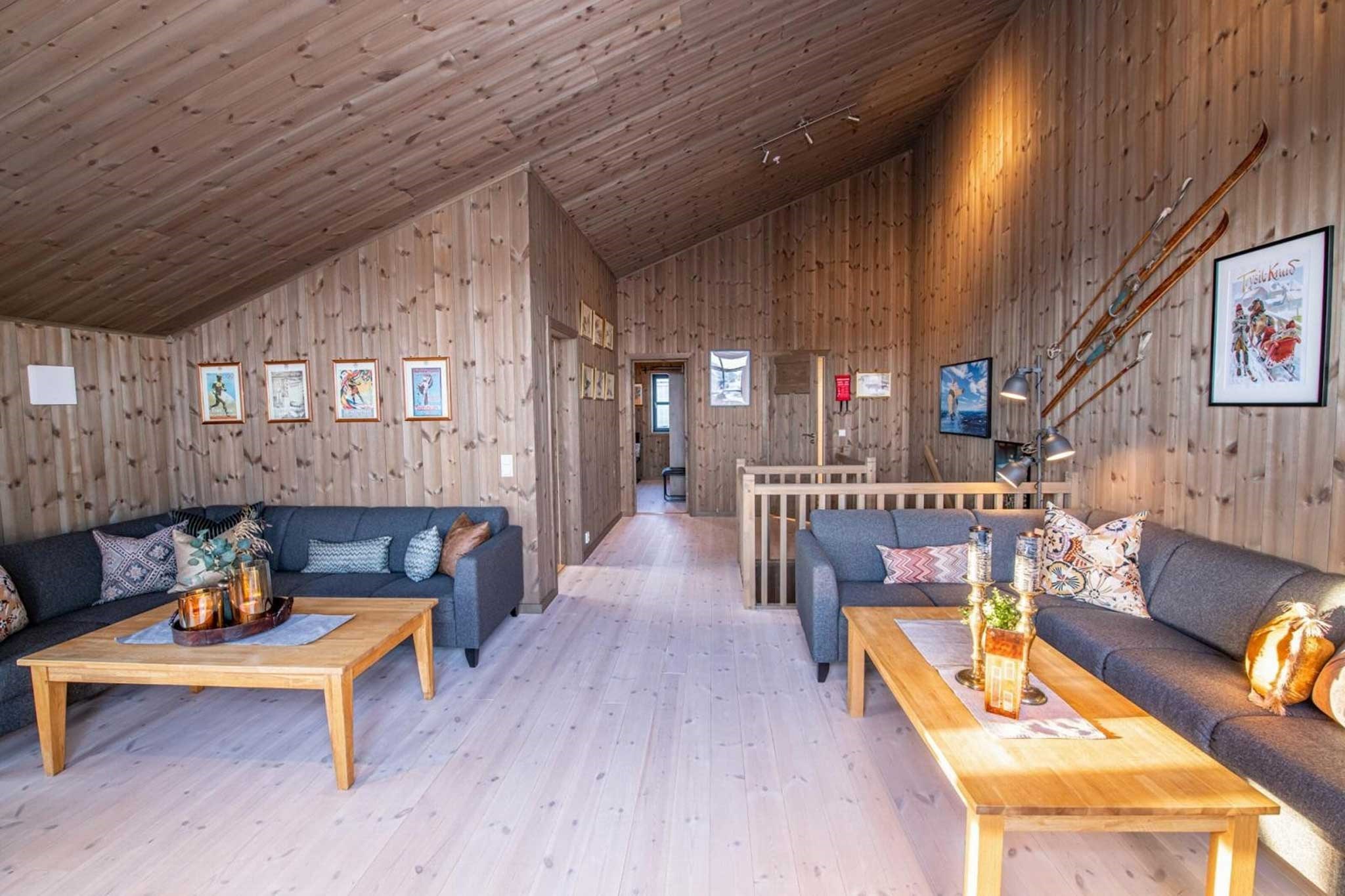 En hyggelig stue med trægulv og -vægge, to mørke sofaer med mønstrede puder, to sofaborde i træ, vægkunst, vintageski på væggen og varmt sollys, der fylder rummet.