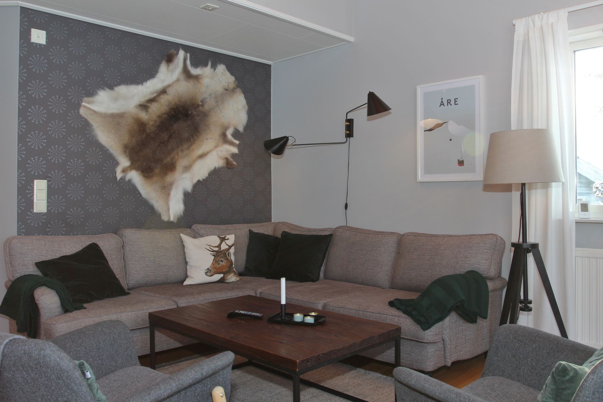 En hyggelig stue med en grå hjørnesofa, mørkegrønne hynder, et sofabord i træ, vægmonterede lamper, et pelsdække på væggen og et vindue med hvide gardiner, der lukker naturligt lys ind.