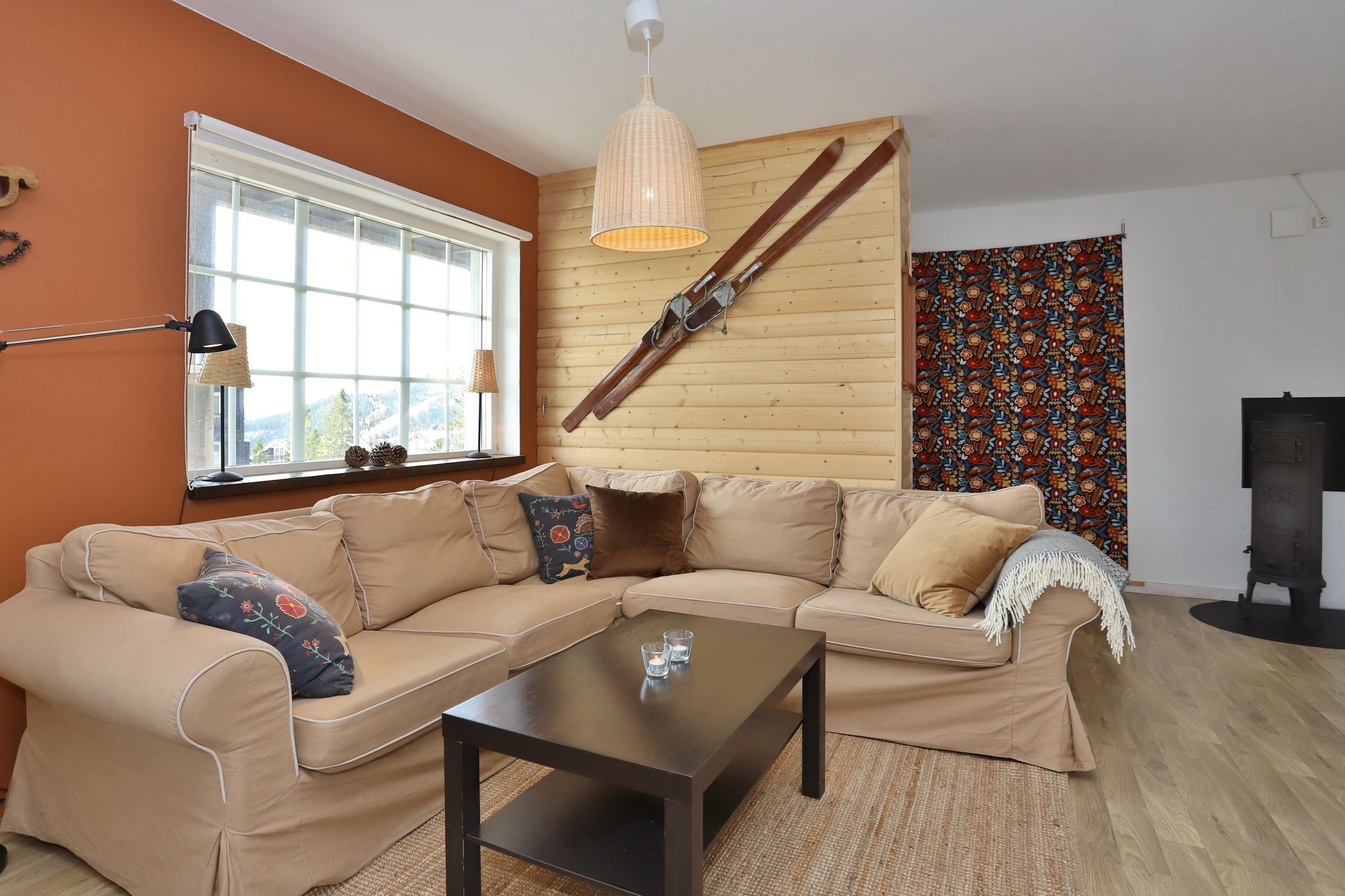 En hyggelig stue med en beige hjørnesofa, farverige puder, et mørkt sofabord og et stort vindue. En trævæg med vintageski pryder rummet sammen med et blomstret gardin og en sort brændeovn.