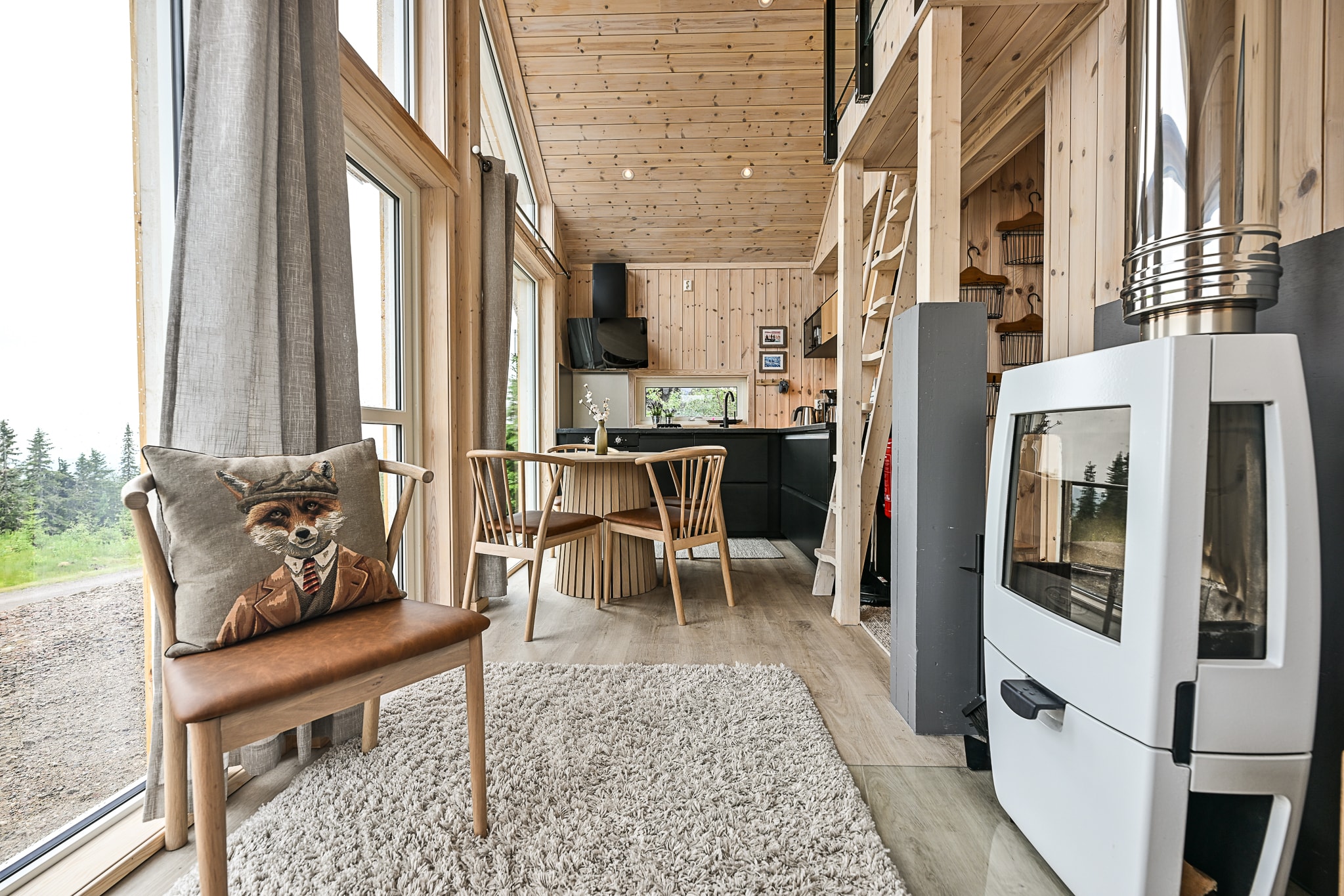 Lyst, moderne lille husindretning med trævægge, store vinduer, et hyggeligt tæppe, en brændeovn, en stol med en rævepude, et spisebord og en stige, der fører op til en hems.