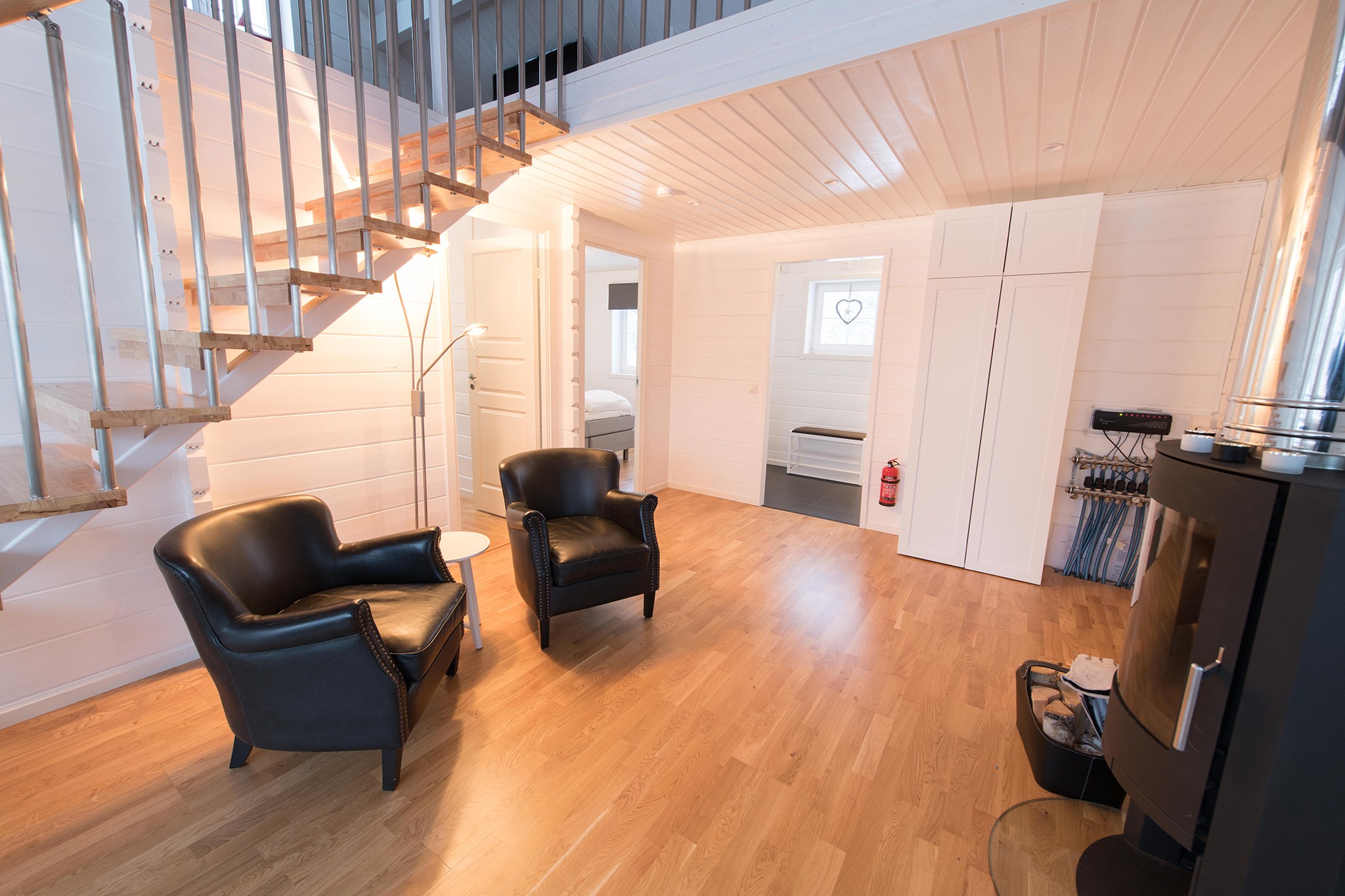 Moderne, lyst opholdsrum med trægulve, to sorte lænestole, en trappe med metalgelændere, en brændeovn og døre, der fører til andre rum. En lampe, brandslukker og opbevaringsområde er også synlige.