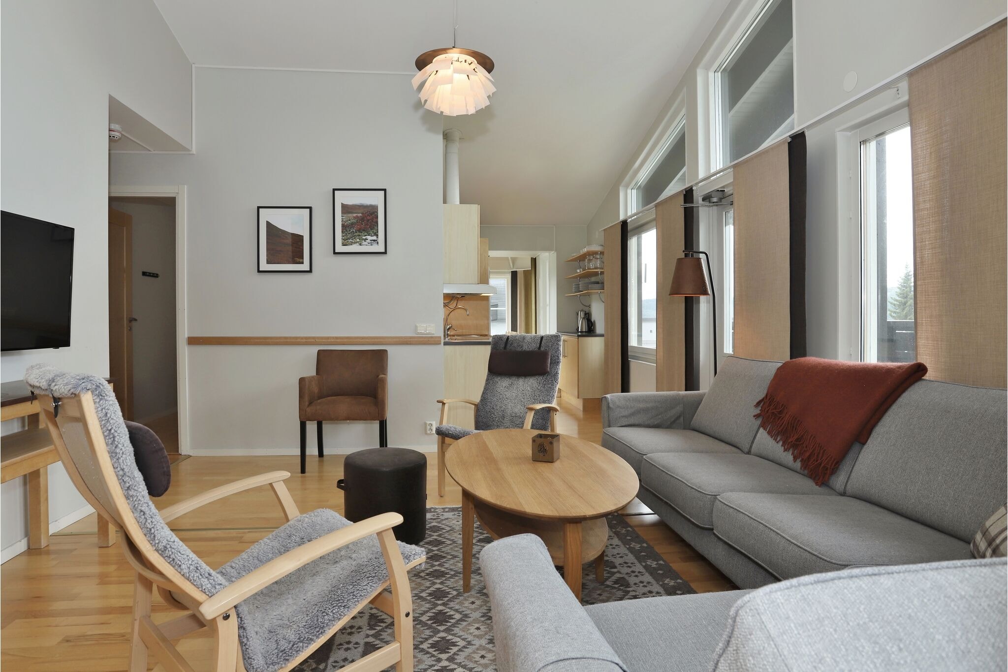 Moderne stue med lyse trægulve, grå sofa, to lænestole, et rundt sofabord i træ, vægkunst og store vinduer med neutrale persienner. Værelset har et hyggeligt, skandinavisk-inspireret design.