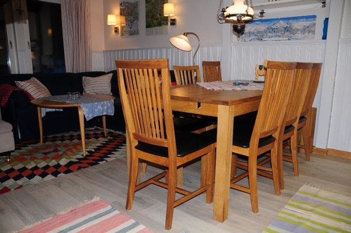 En hyggelig spiseplads med et træbord og seks matchende stole, en mørk sofa med stribede puder, farverige tæpper, et rundt sidebord og vægkunst under varm belysning.