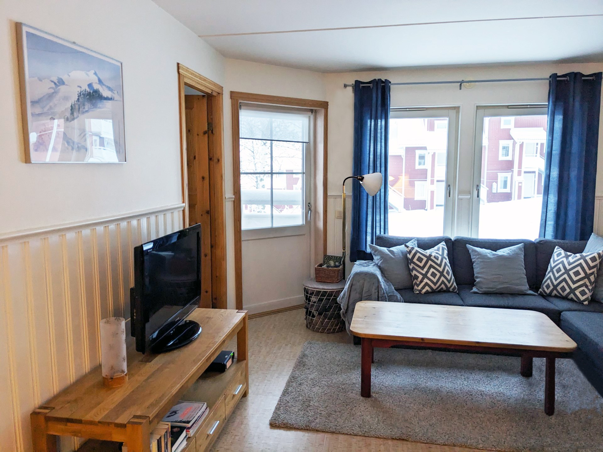 En hyggelig stue med en grå hjørnesofa, mønstrede puder, et sofabord i træ, et tv på en træstander, et vindue med marineblå gardiner og synlige sneklædte bygninger udenfor. Et bjergfoto hænger på væggen.