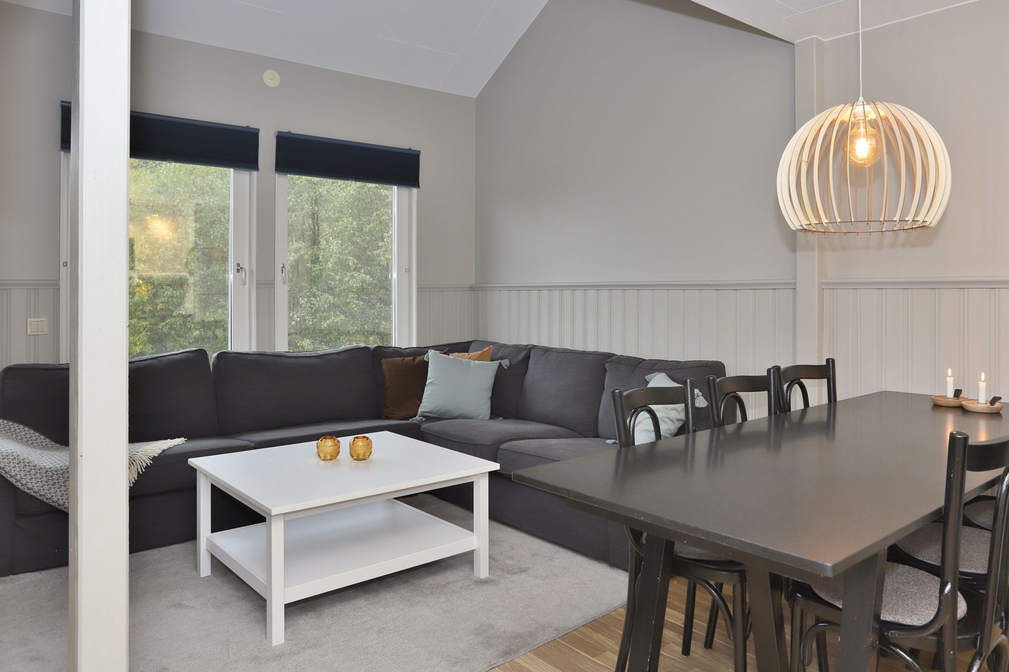 En moderne opholds- og spisestue med en mørkegrå sofa, et hvidt sofabord, et sort spisebord med stole og en stor træpendellampe; to vinduer lukker naturligt lys ind og fremhæver neutrale toner overalt.