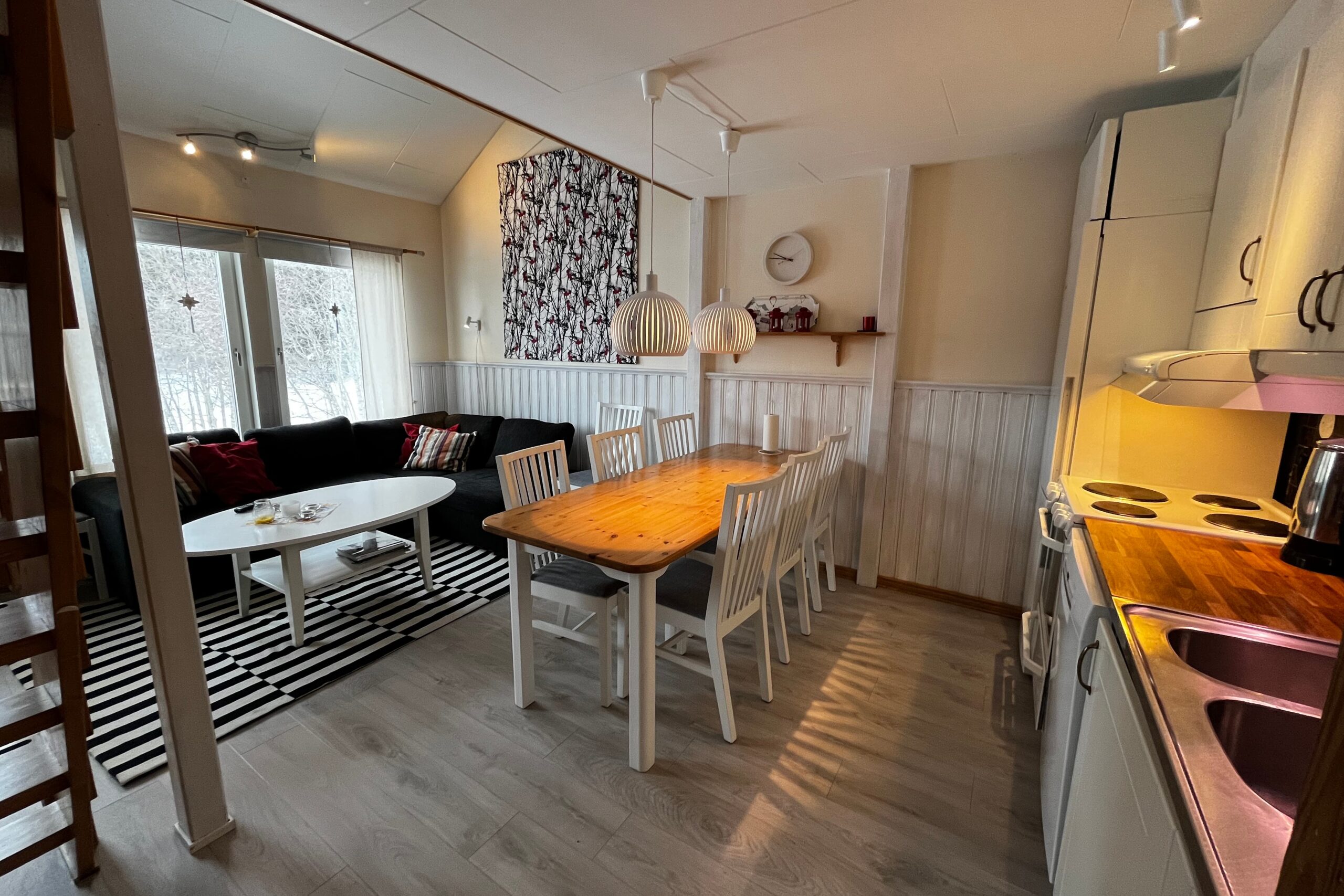 En hyggelig åben stue og spisestue med et træbord, seks hvide stole, en sort sofa, stribet tæppe, sofabord og et lille køkken med hvide skabe. Store vinduer viser et snedækket landskab udenfor.