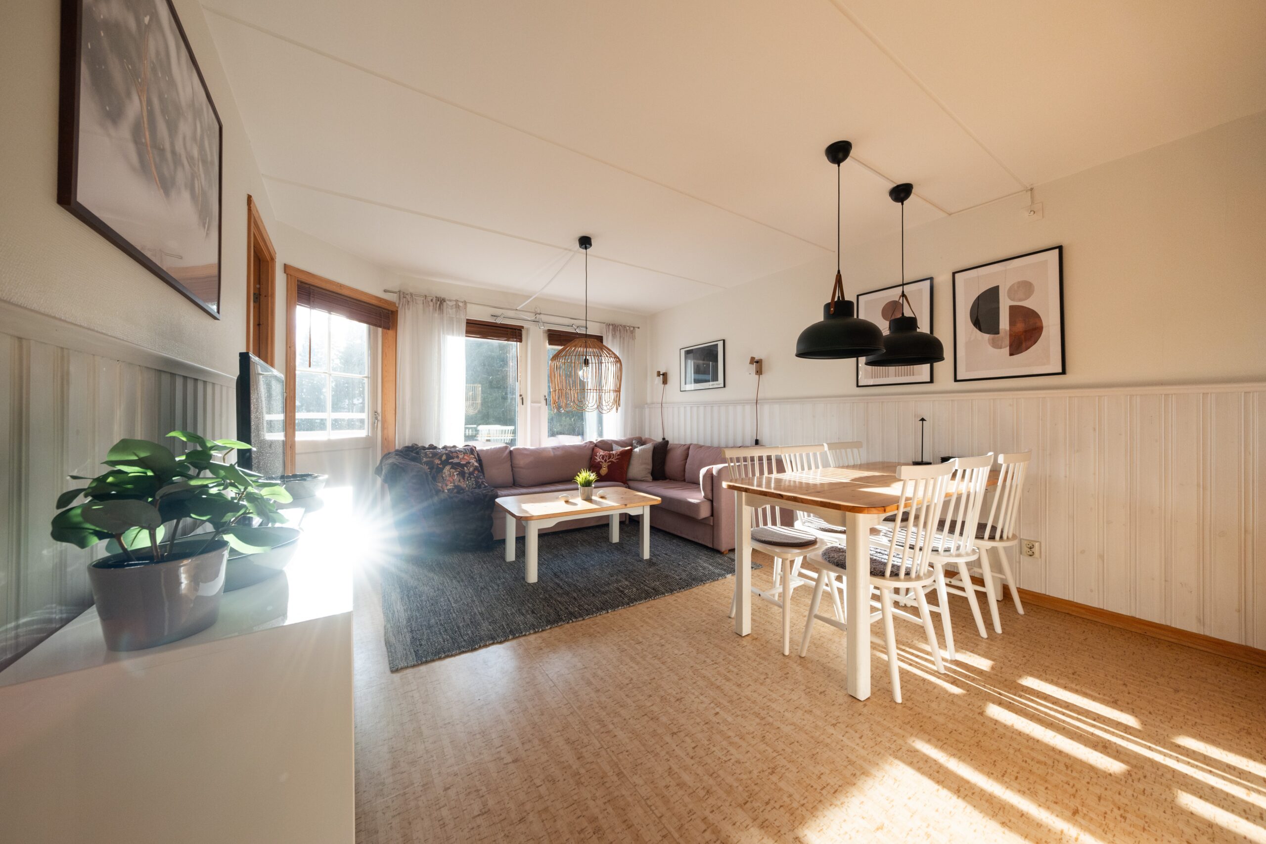 Lys, moderne opholds- og spisestue med trægulve, hvide vægge, et lyst træspisebord med hvide stole, en hyggelig sofa, sofabord, store vinduer, der lukker sollyset ind, og vægkunst.