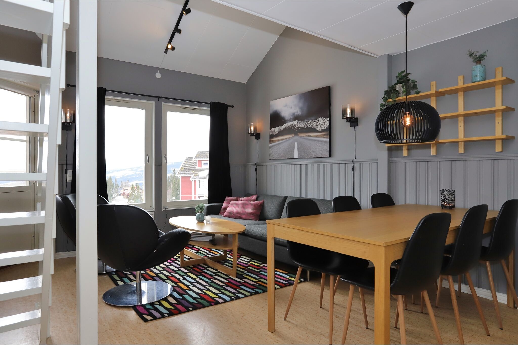 Moderne opholds- og spisestue med grå vægge, en sofa, sorte stole, et spisebord i træ og et rundt sofabord. Store vinduer lukker naturligt lys ind. Et farverigt tæppe og vægkunst tilføjer visuel interesse til rummet.