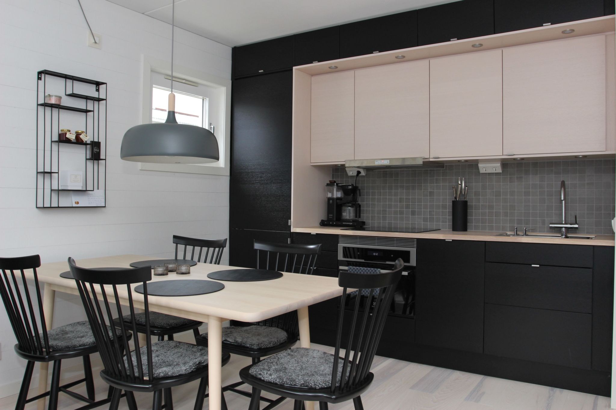 Moderne køkken med sorte skabe, et lyst træ spisebordssæt til fire med sorte stole, en grå pendellampe ovenover, et lille vindue, flisebelagt bagplade og en væghylde med indretningsartikler. Rummet er rent og minimalistisk.