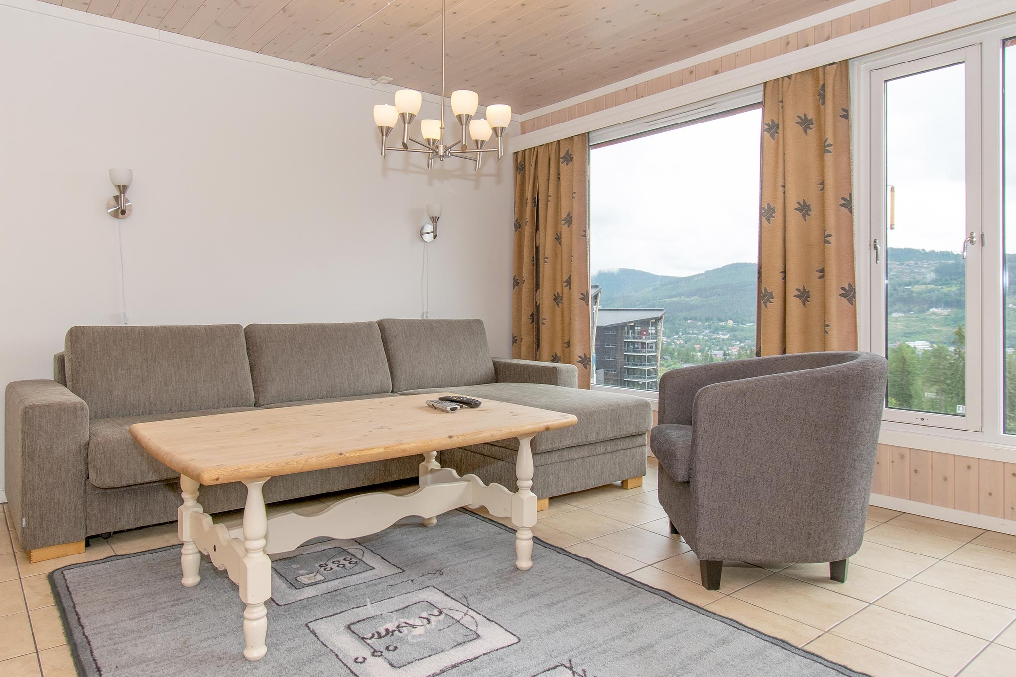 En lys stue med en grå sofa, et sofabord i lyst træ, en grå lænestol, mønstrede gardiner, et stort vindue med en smuk udsigt over bjergene og en moderne lysekrone ovenover.