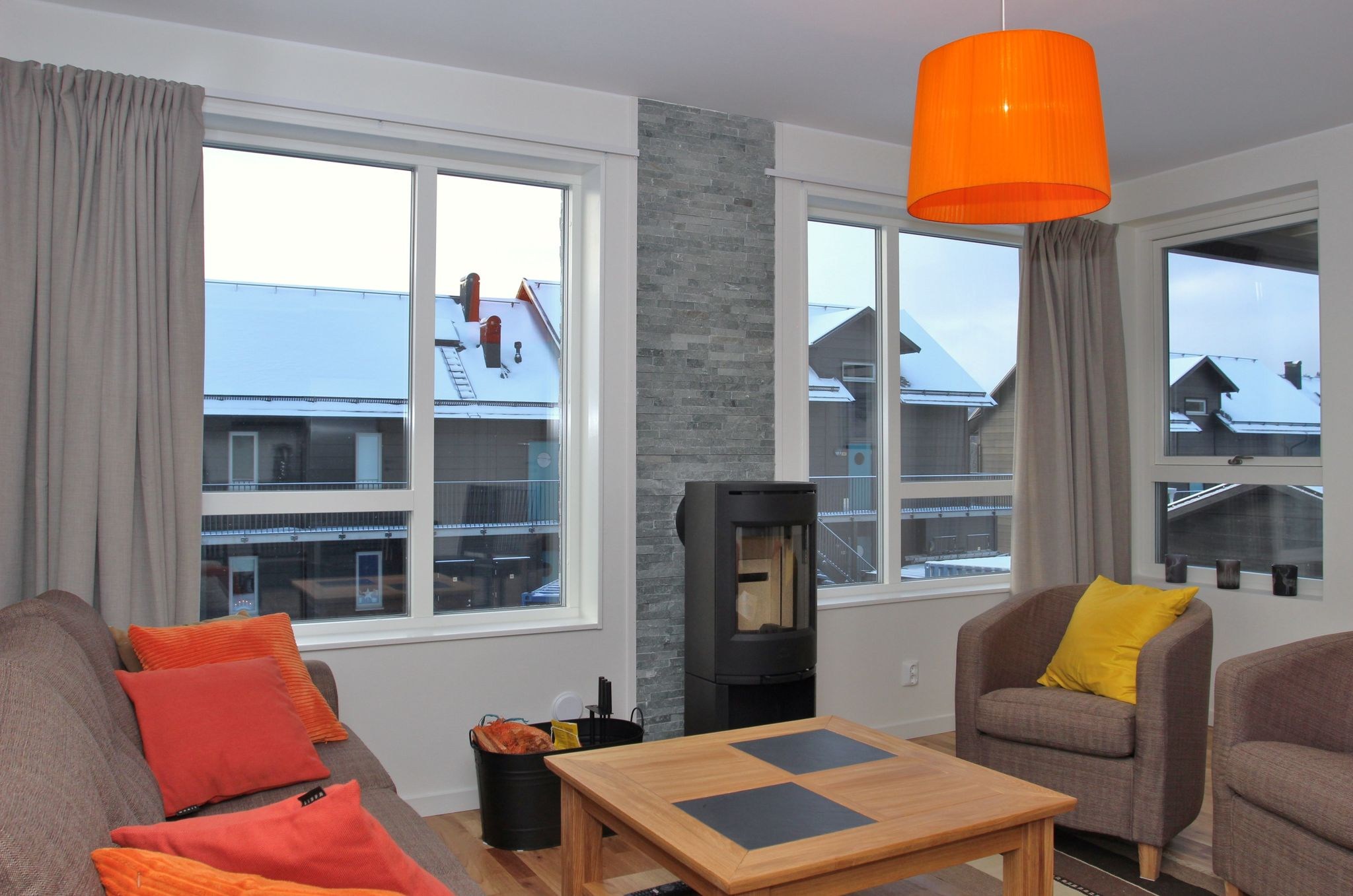 En hyggelig stue med store vinduer, en brun sofa og en grå lænestol med farverige hynder, en brændeovn, et sofabord i træ og en orange hængelampe. Snedækkede hustage er synlige udenfor.